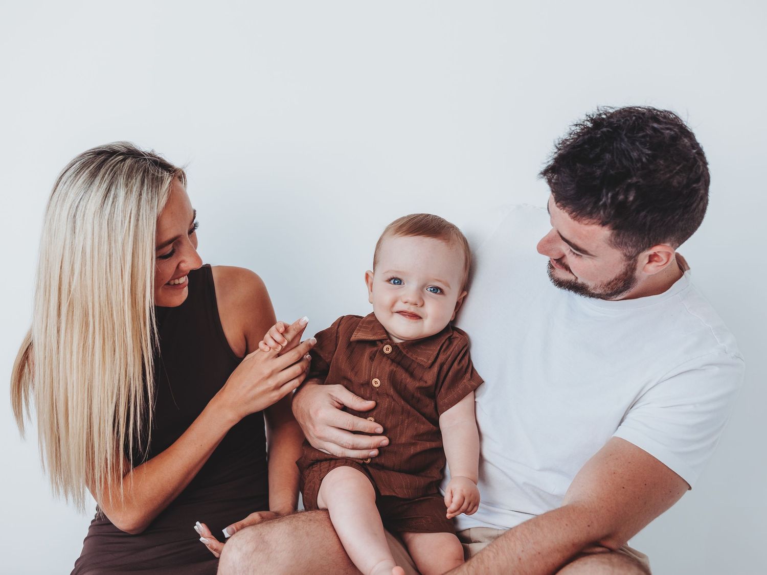 Happy young family with blonde mother, bearded father, and smiling baby in brown outfit sitting together.