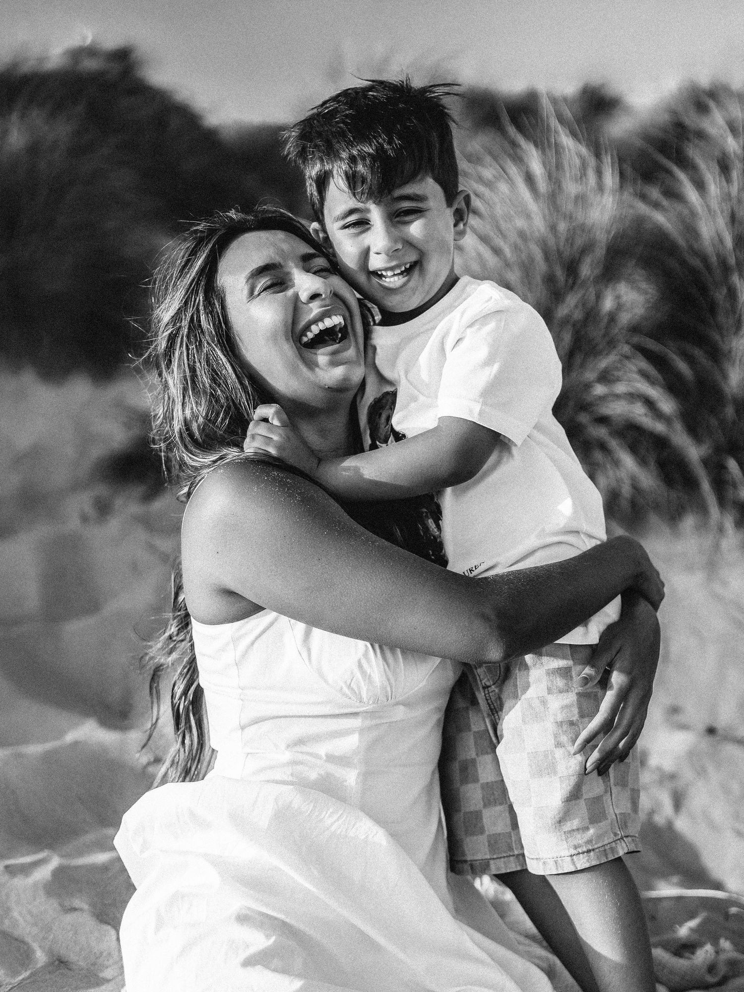 Black and white photo of a laughing woman in white dress hugging a smiling boy on a sandy beach.