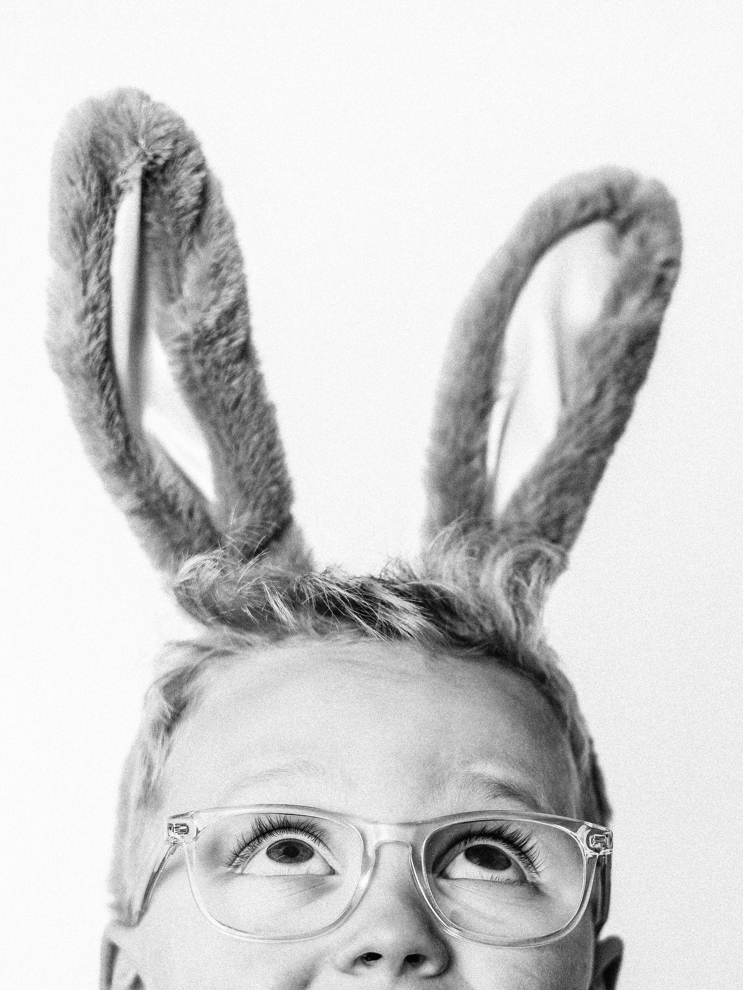 Black and white photo of a child wearing fuzzy bunny ears headband and clear glasses, looking upward with wide eyes.