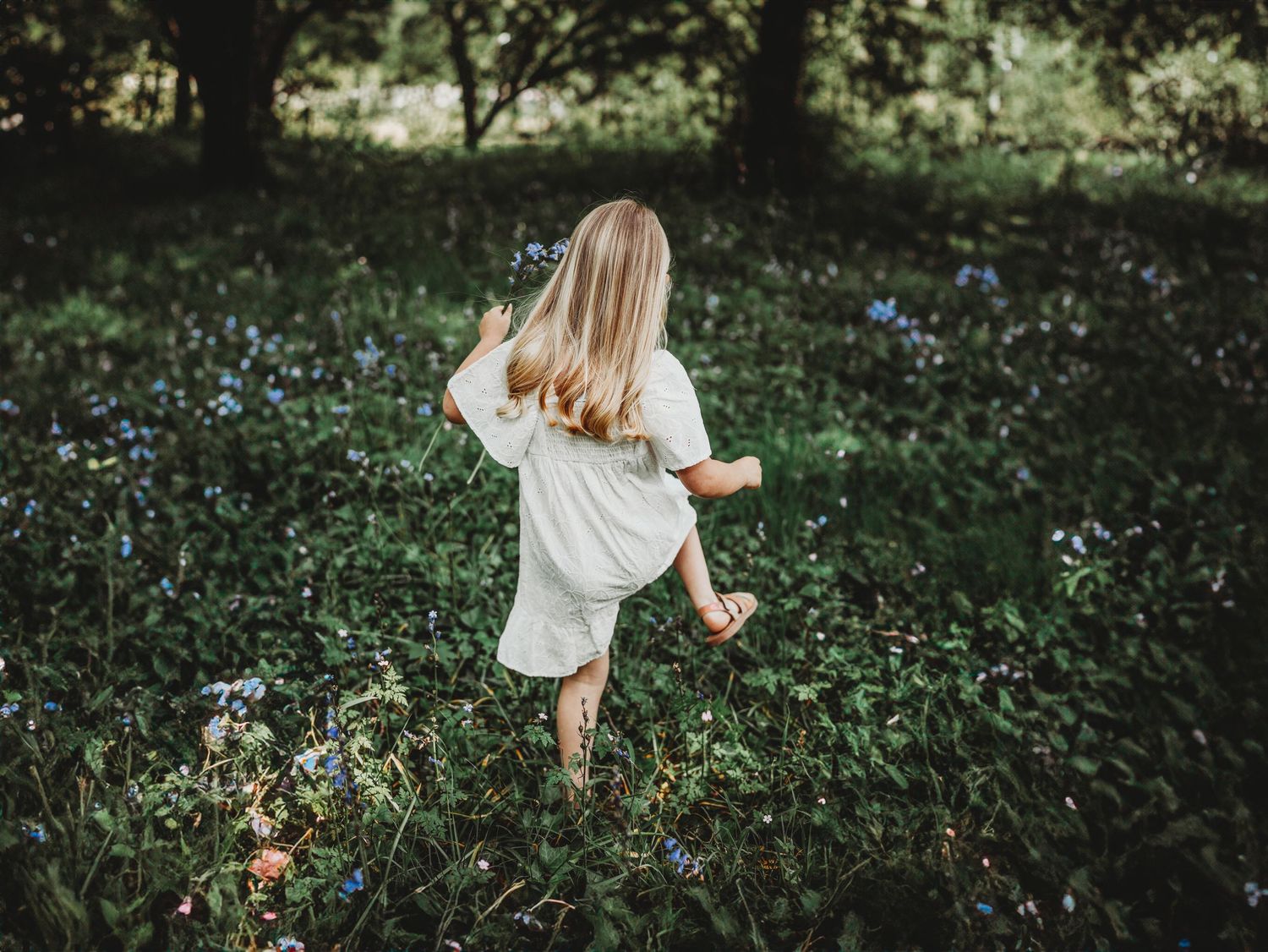 Young girl in white dress walking barefoot through lush green field with blue wildflowers in forest.