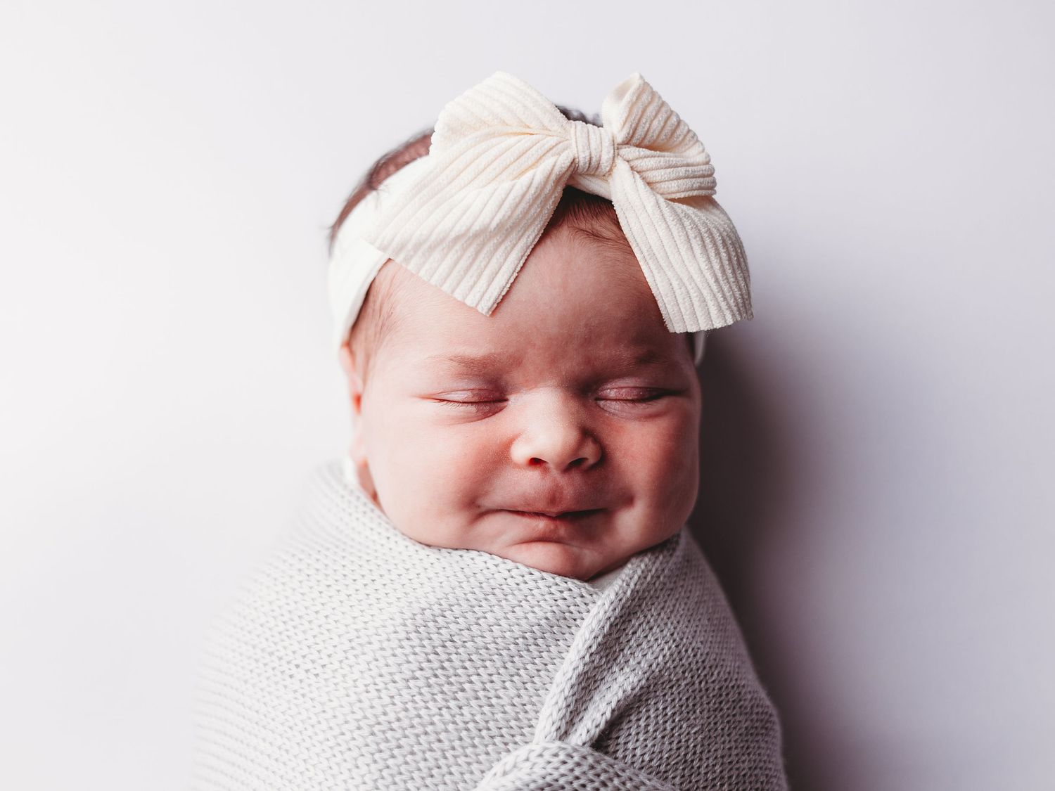A newborn baby wrapped in white swaddle with a striped bow headband smiles while sleeping against a light background.