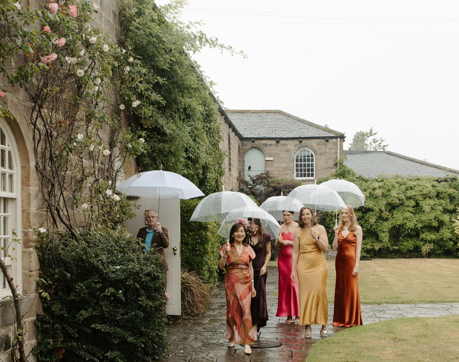 Wedding party stands under white umbrellas beside a historic stone building on a cloudy day.