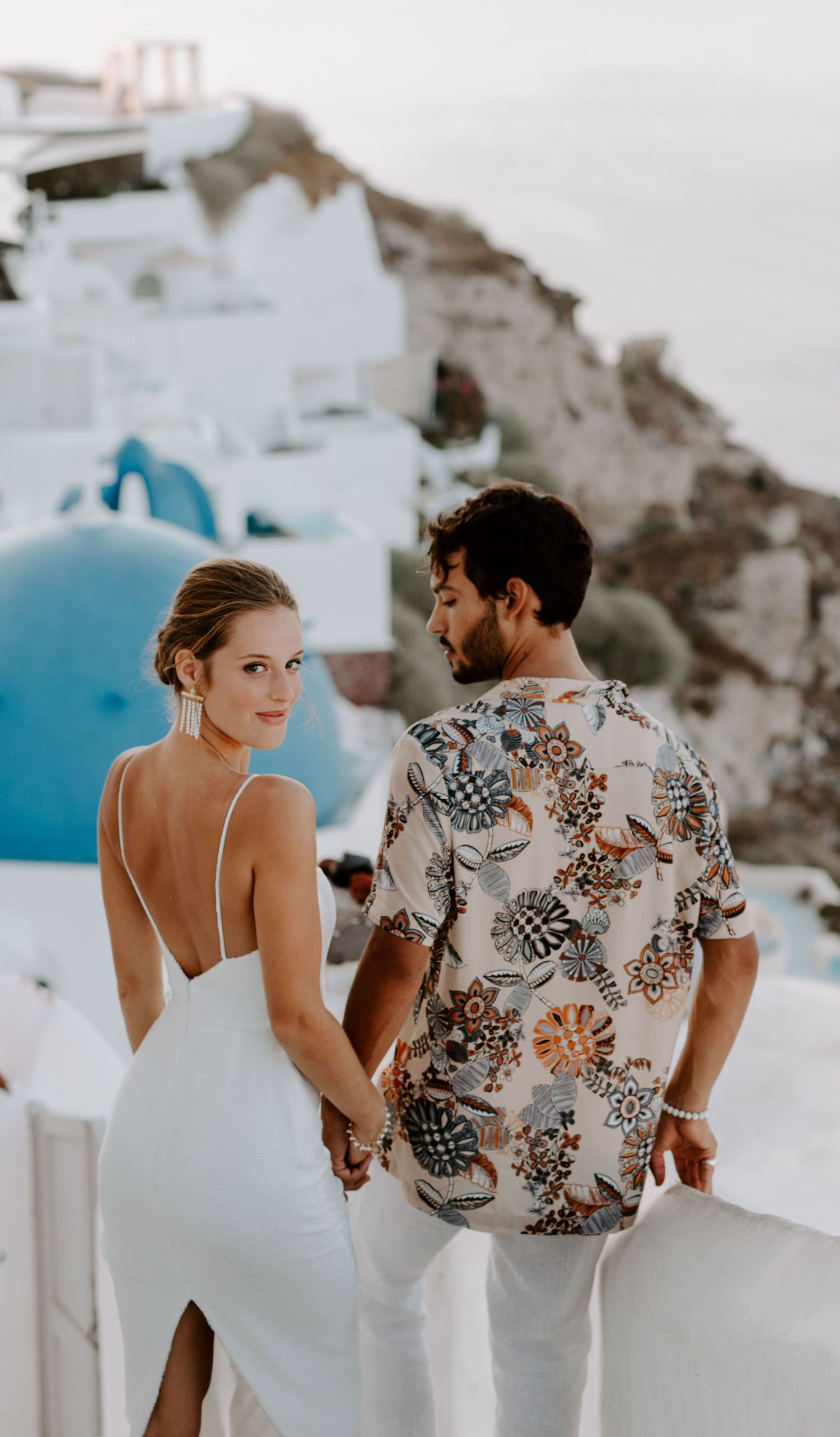 Couple in white and floral attire standing on a Mediterranean terrace overlooking the sea.