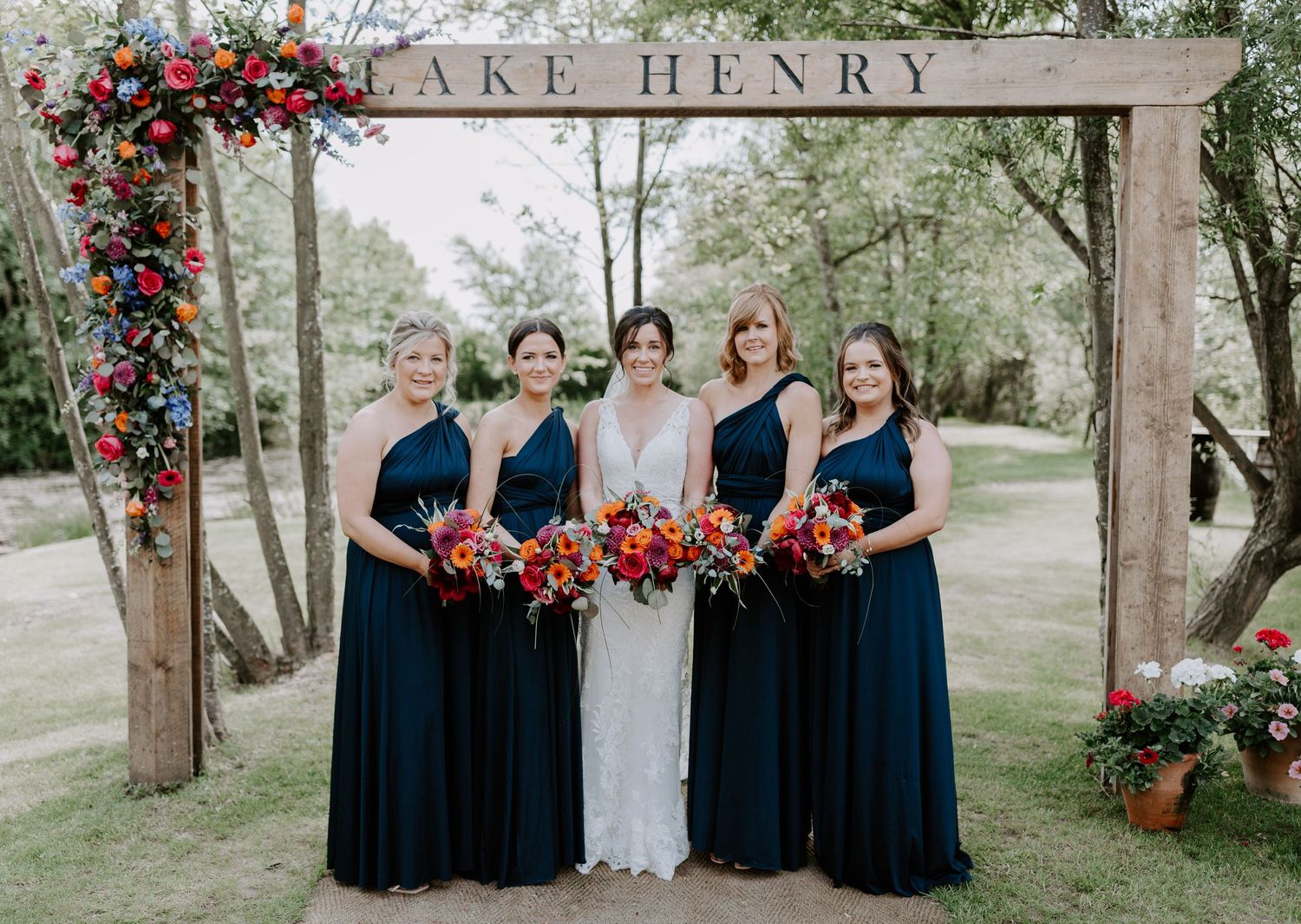 Wedding party in navy blue dresses posing under a wooden arch with red floral decorations.