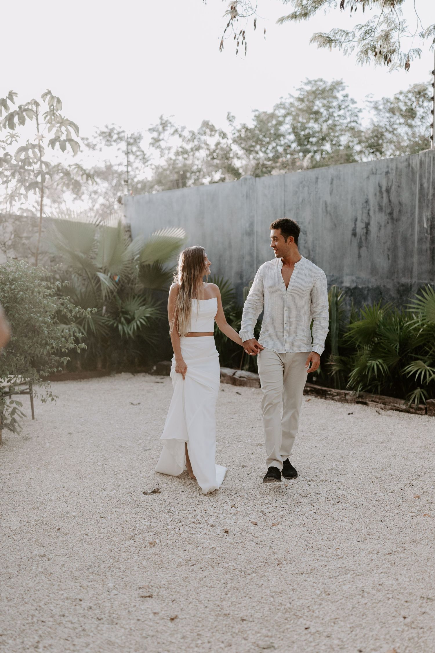 A couple walks together on a sandy path surrounded by tropical landscaping.