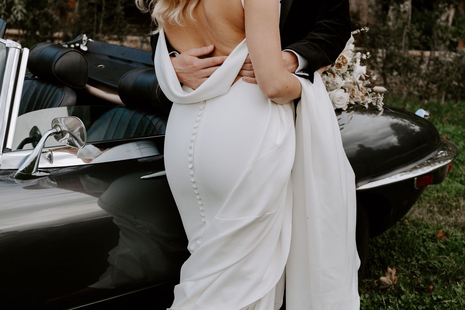 A classic car parked outdoors with a white-clad couple posing beside it.