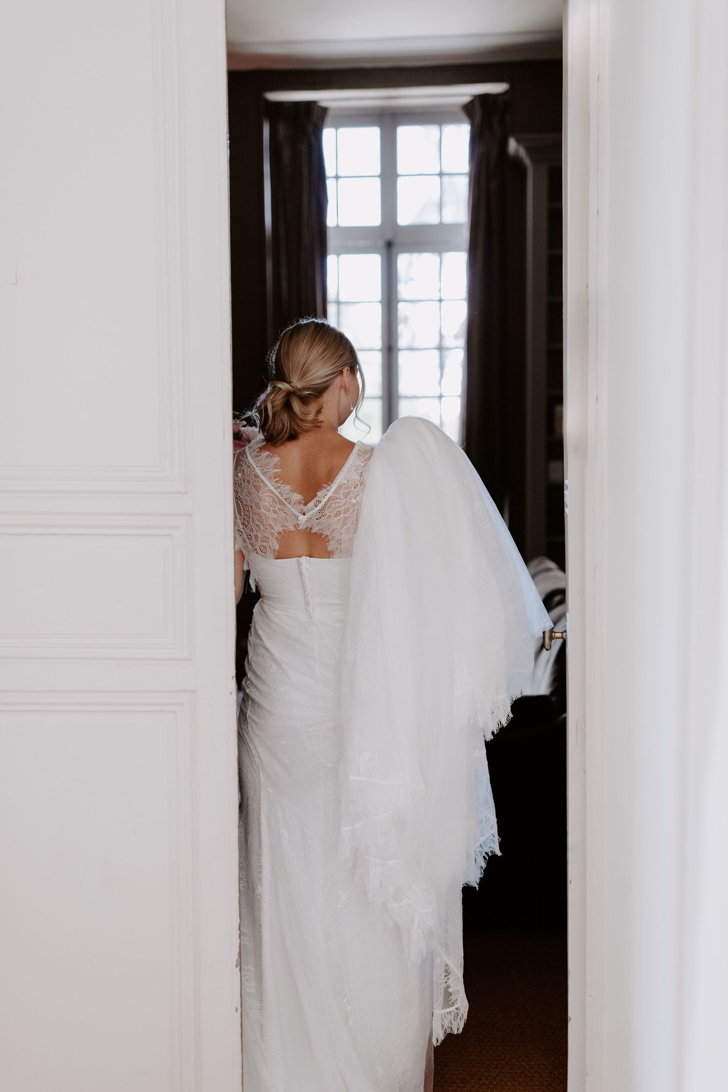 A bride in a flowing white wedding gown stands elegantly in a doorway with natural light streaming in.