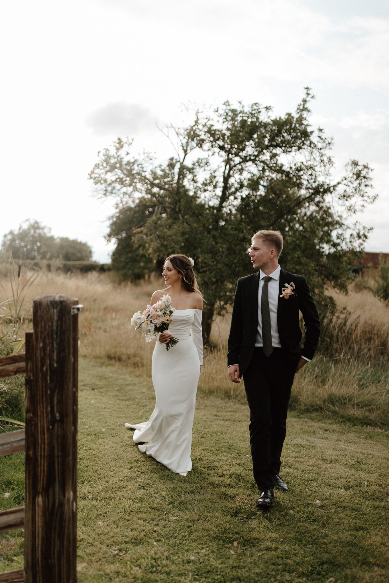 Newlyweds walking together on grassy hill with rustic wooden fence posts.