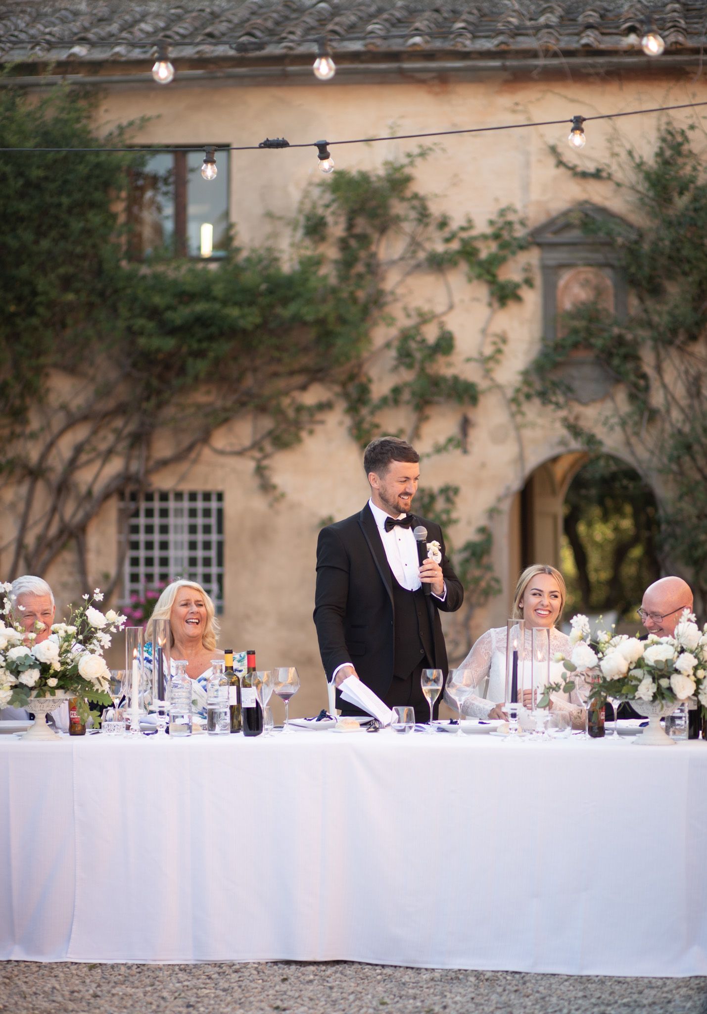 Outdoor wedding reception dinner table with white linens and string lights at dusk.