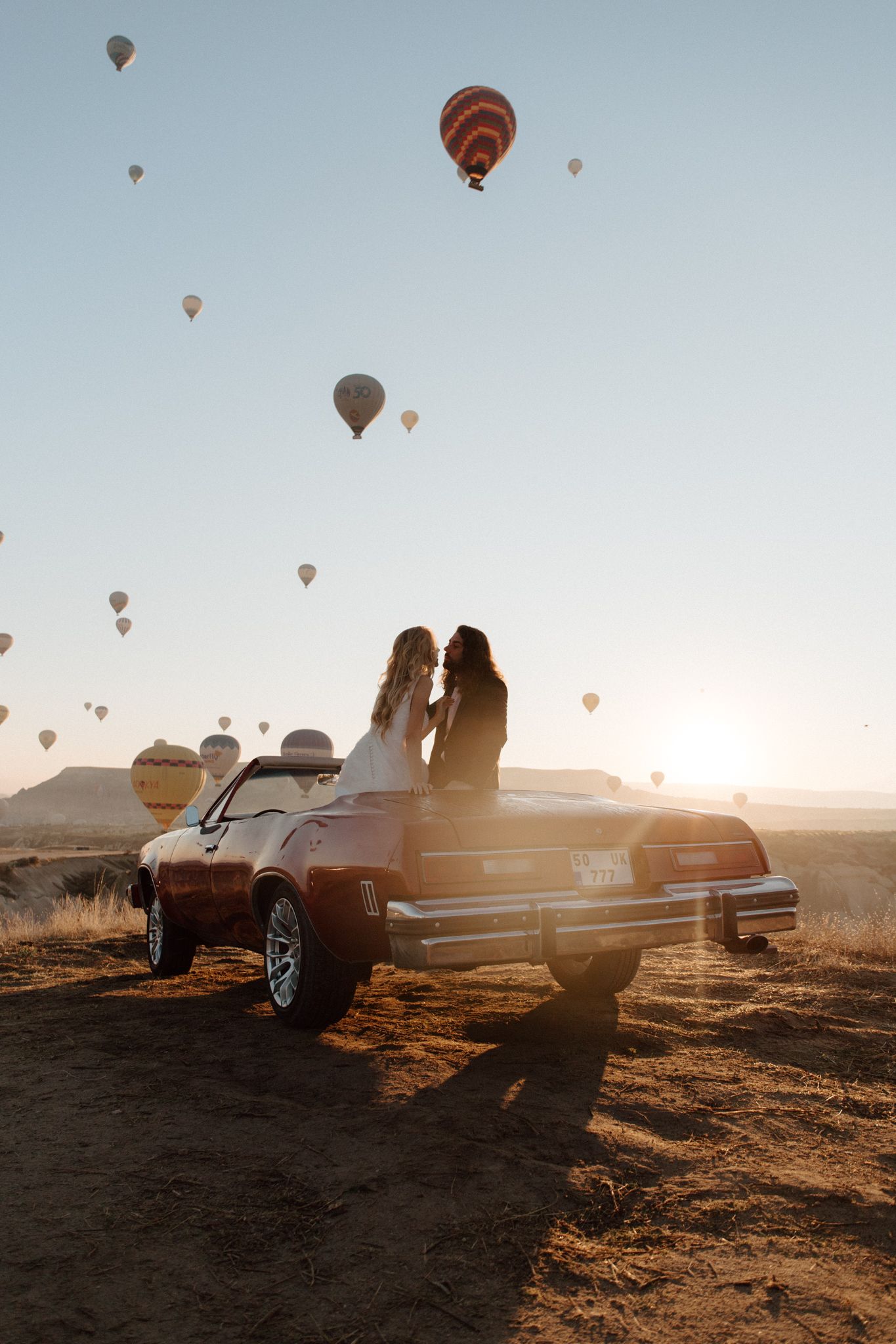 Classic car silhouetted against sunset sky with hot air balloons floating in background.