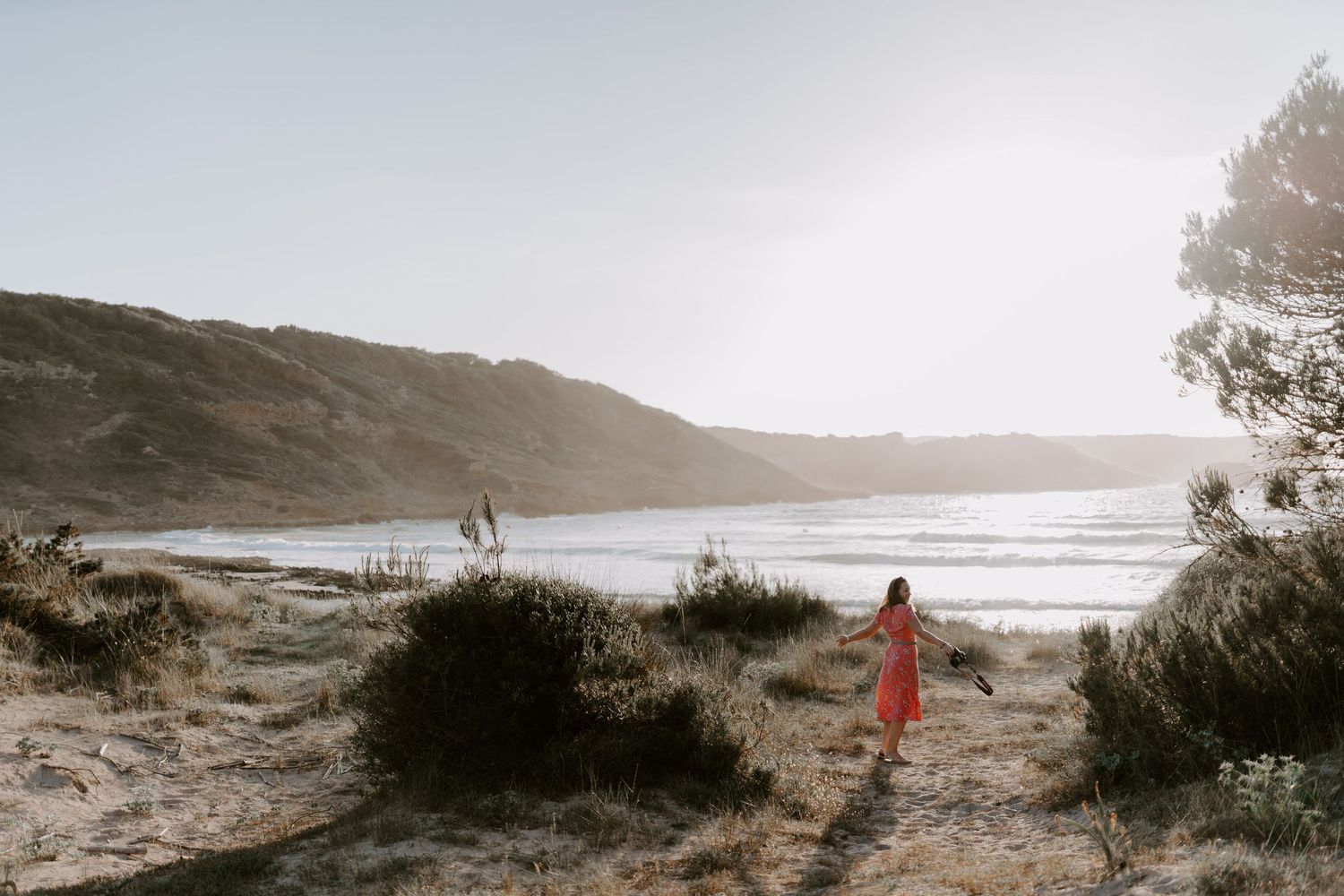 Figure in red dress exploring sandy coastal path during golden hour.