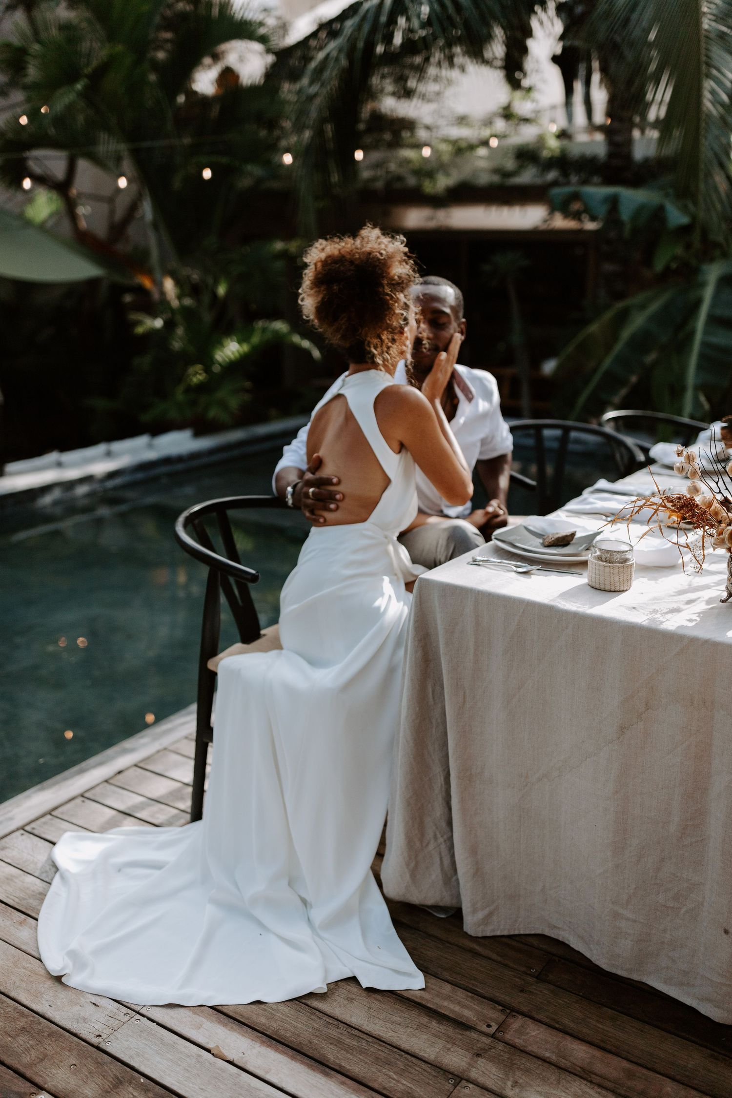 A couple in white attire shares an intimate moment at a rustic outdoor dining table.