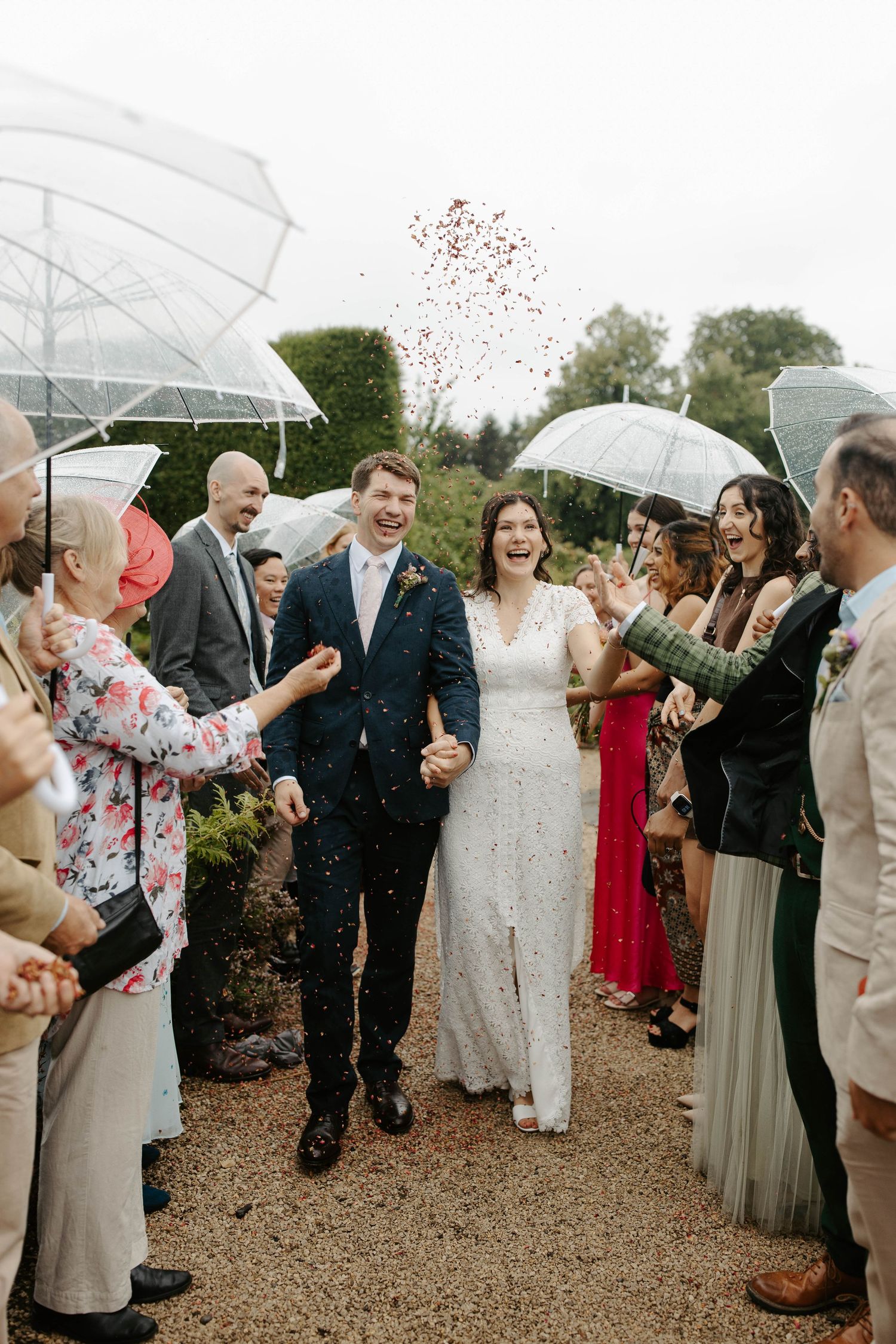 Wedding guests with clear umbrellas throwing confetti during an outdoor celebration.