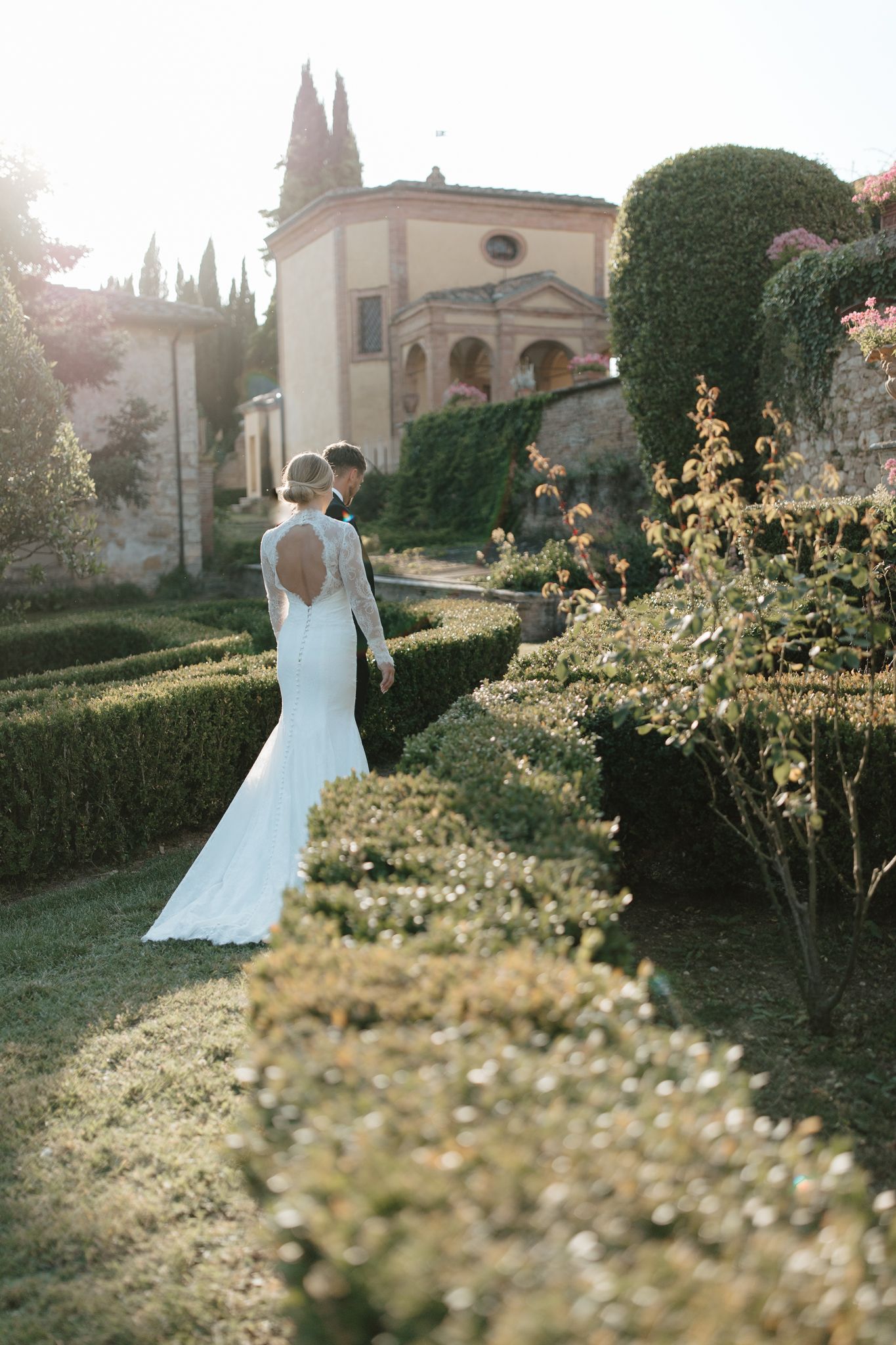 A romantic garden wedding scene at sunset with a flowing white dress among manicured hedges and blooming flowers.