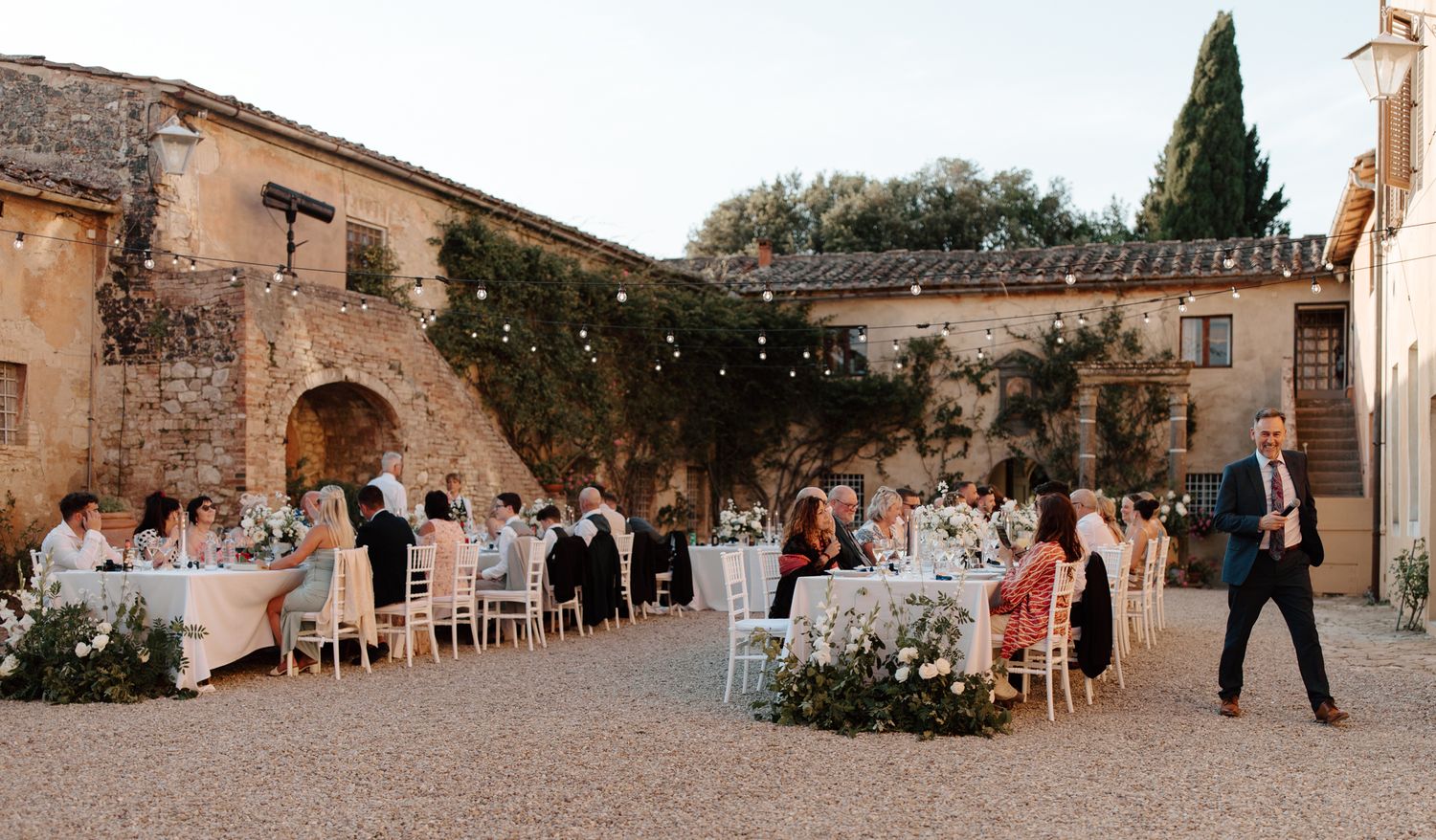Wedding guests dance and celebrate during an outdoor reception at a rustic stone venue with string lights overhead.