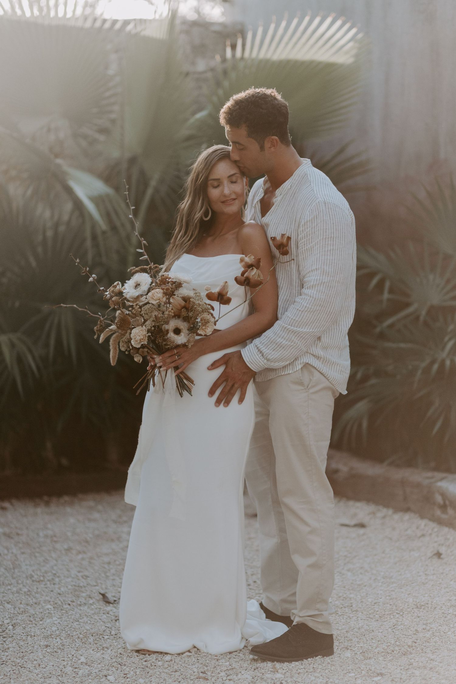 A couple shares an intimate moment surrounded by palm trees and natural lighting.