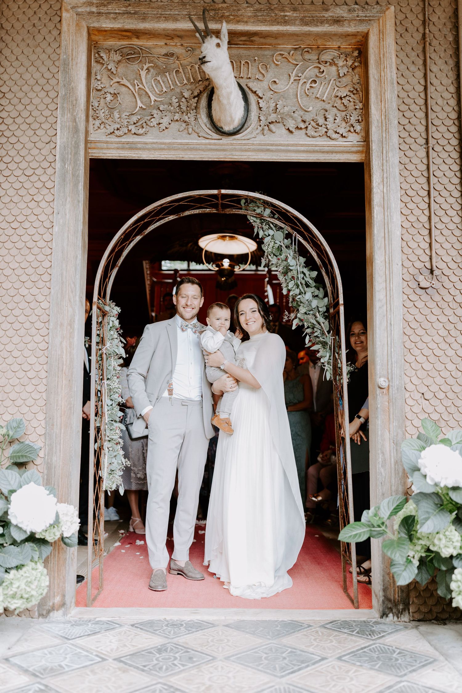 Wedding party members in white attire gather under ornate archways decorated with floral arrangements.