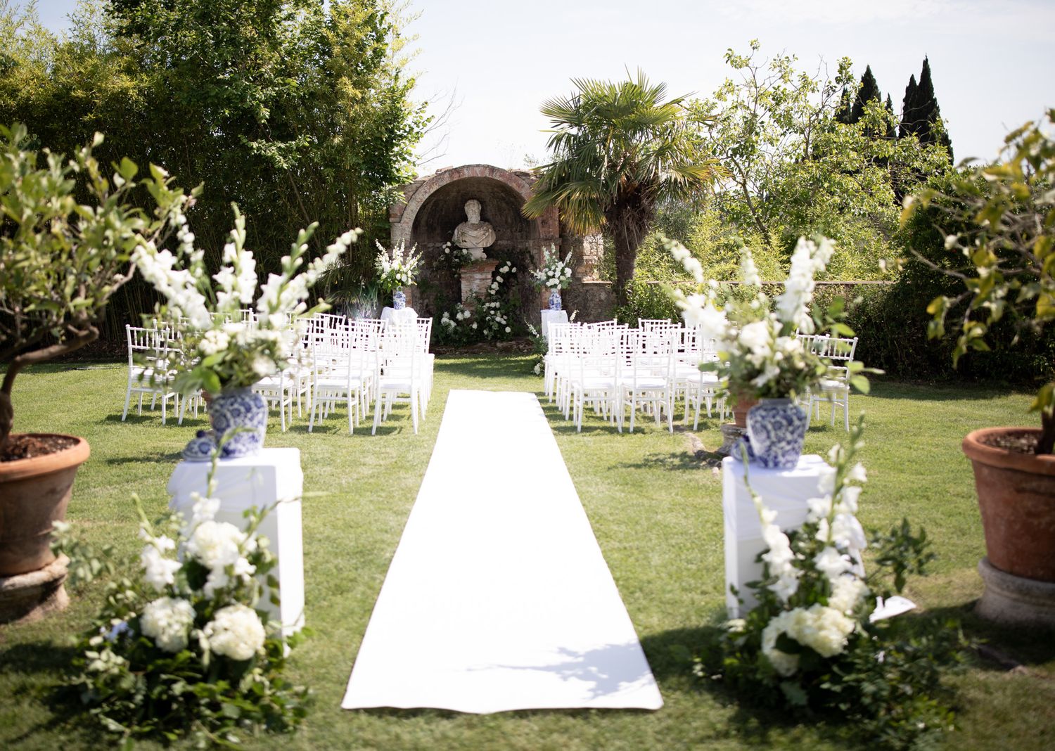 White floral wedding ceremony setup in a lush garden with chairs and decorative arrangements.