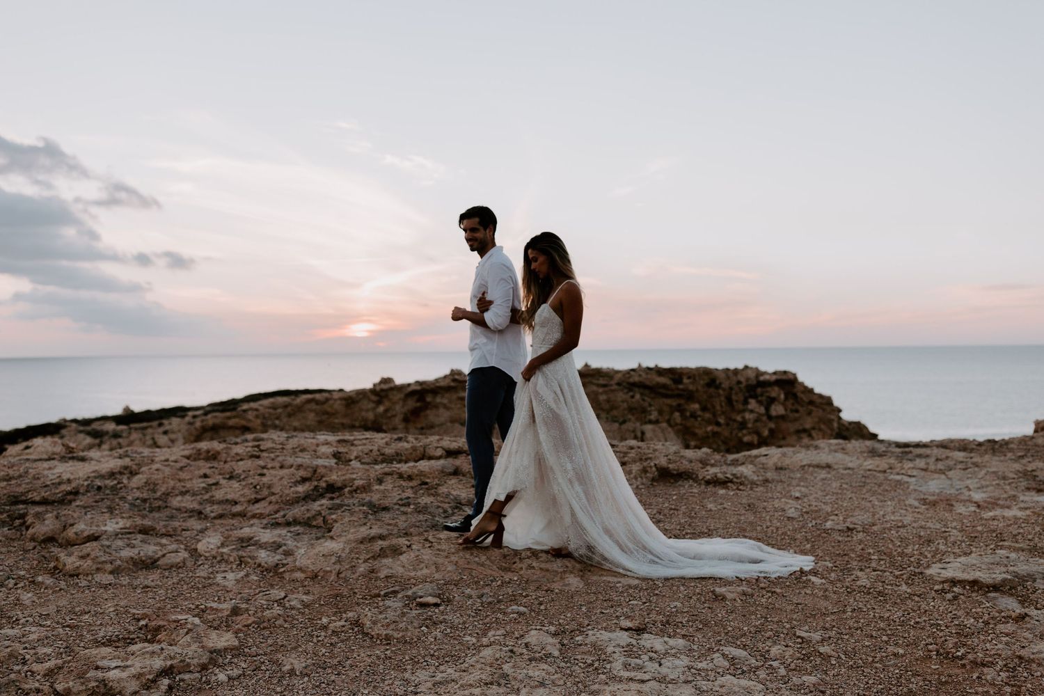 A couple stands on rocky coastal cliffs in Minorca during sunset.