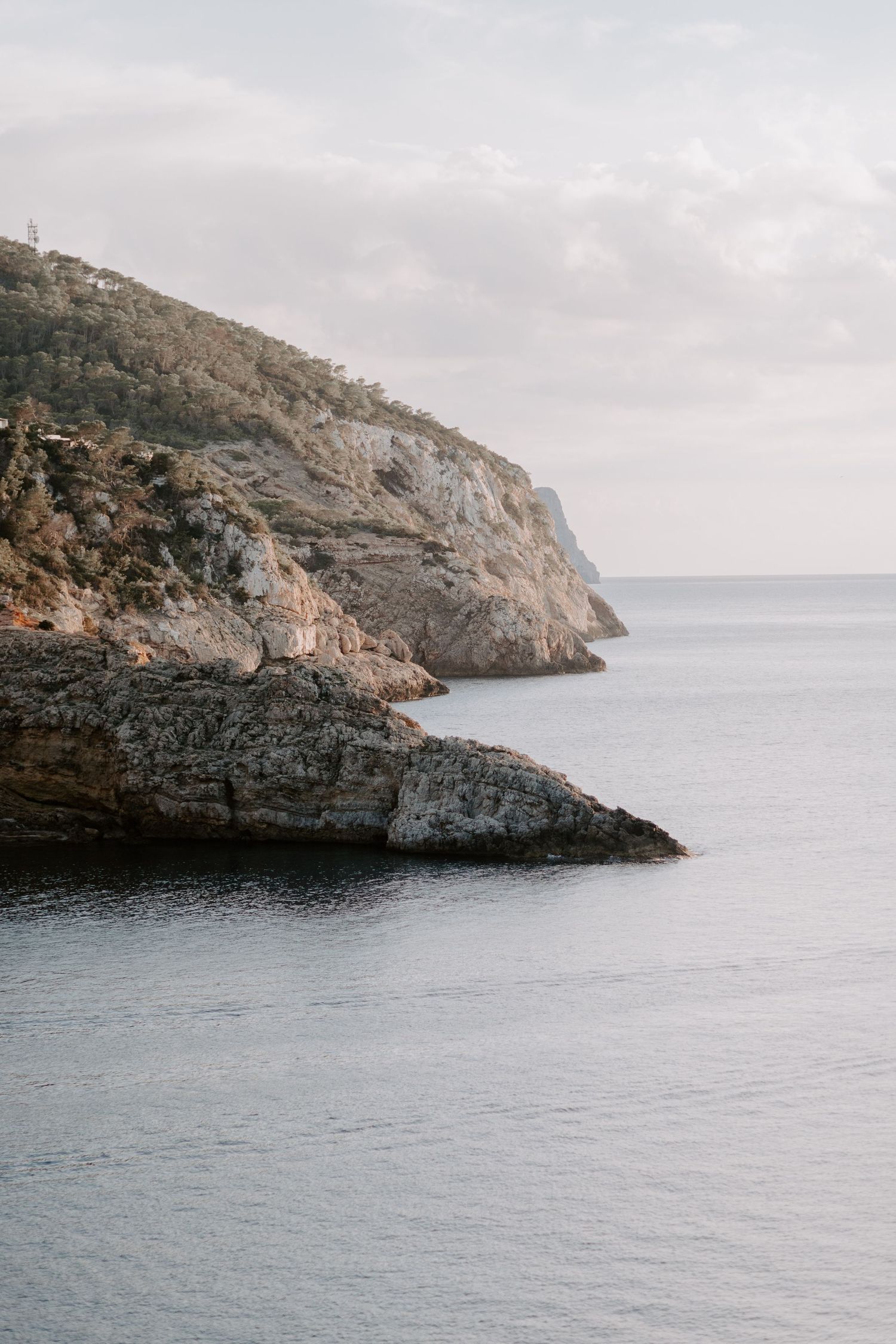 Rocky coastal cliffs meet calm blue waters under an overcast sky along a rugged Mediterranean shoreline.