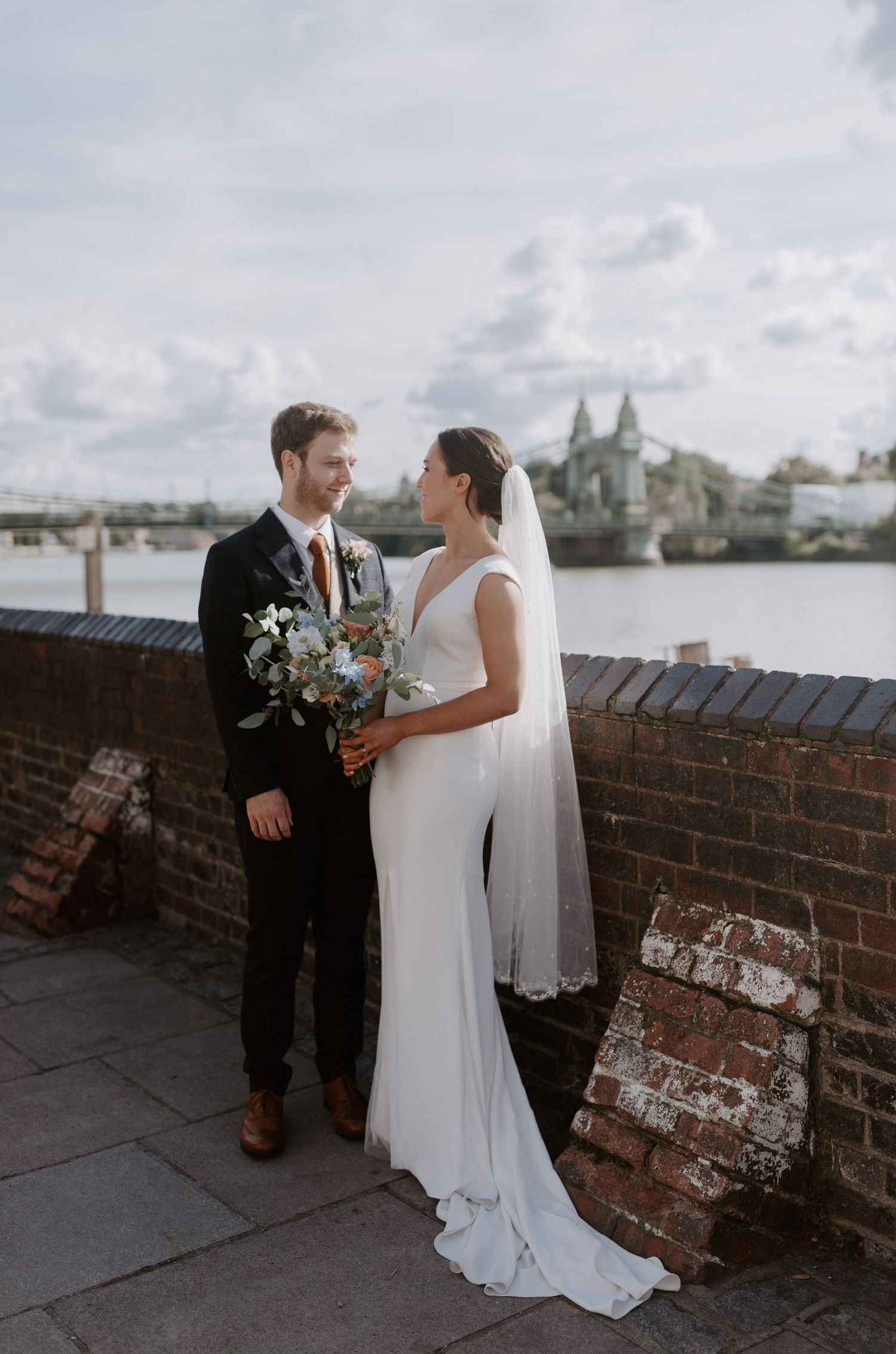 A couple in wedding attire poses against a brick wall with city views.