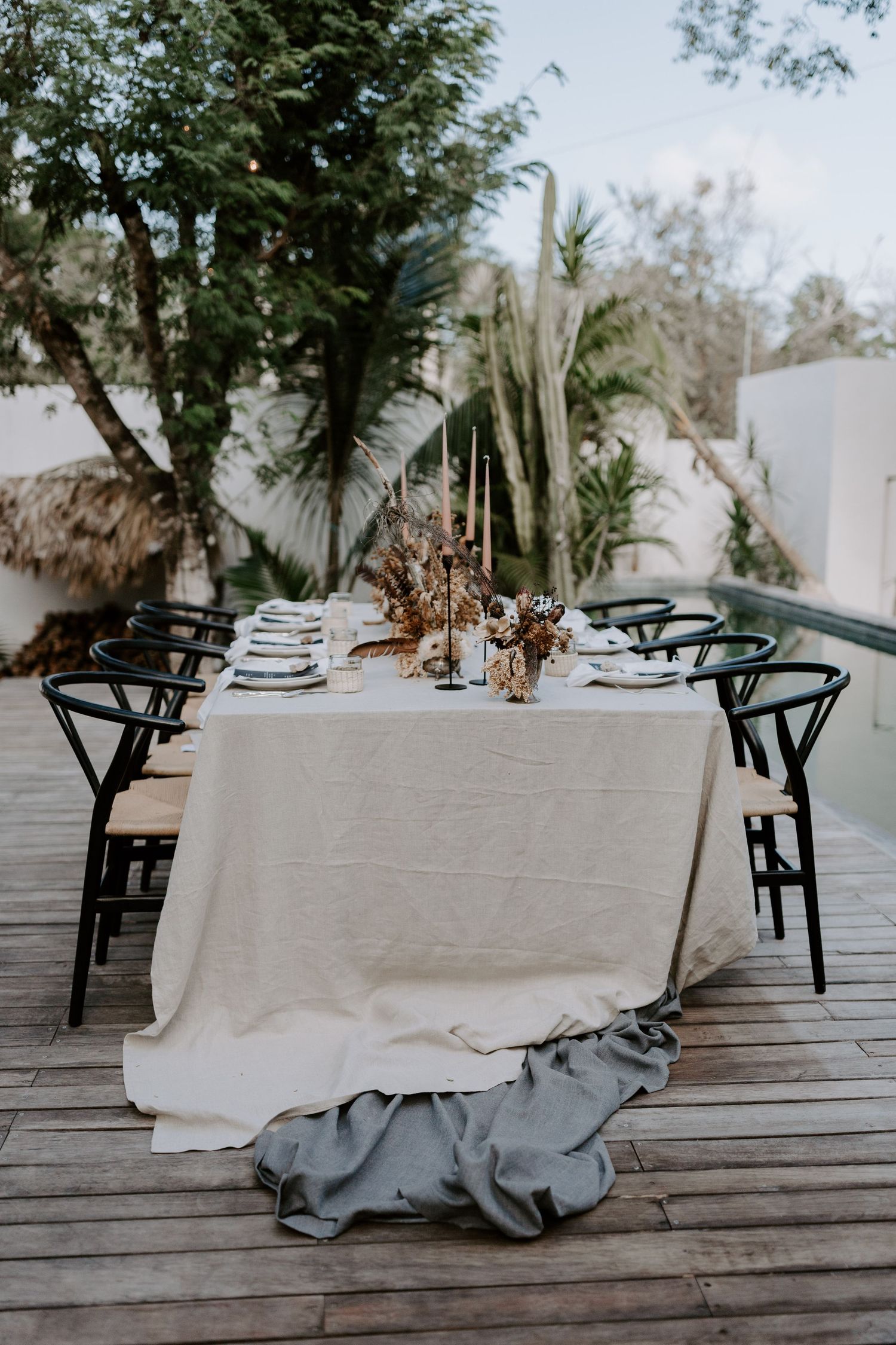 Rustic outdoor dining table setup with linen tablecloth and black chairs on wooden deck.