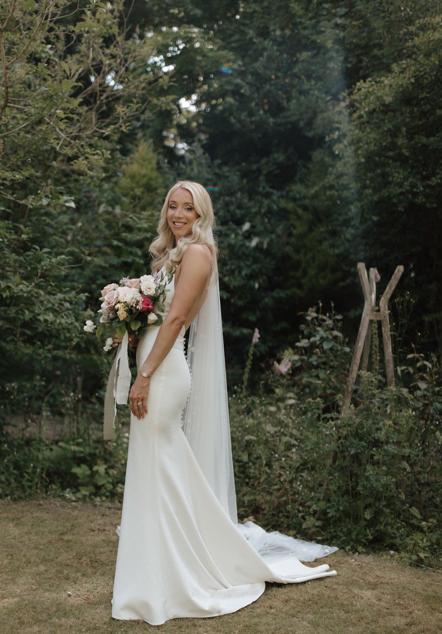 Elegant bride in a sleek white gown holding a romantic bouquet in an outdoor garden setting.