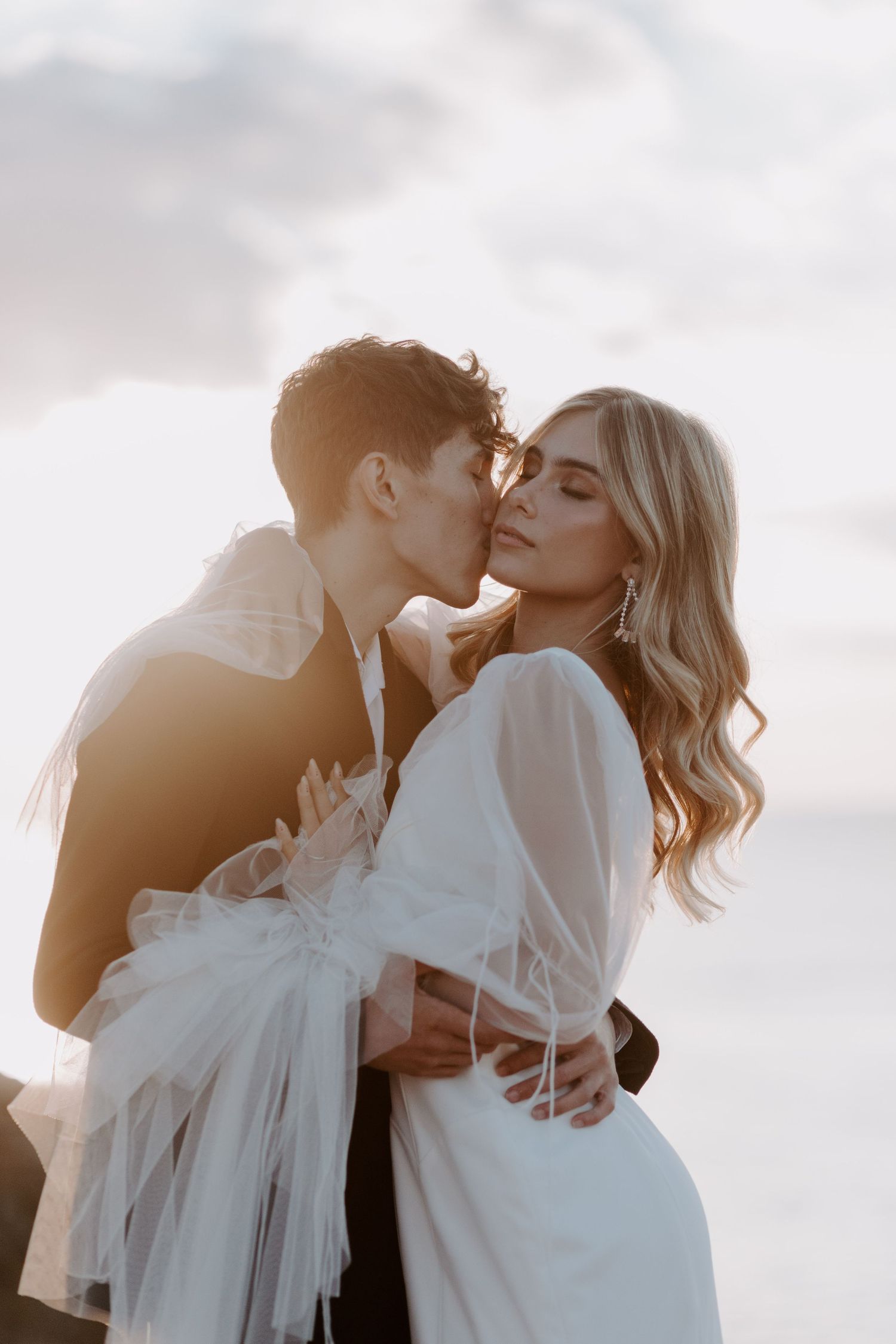 A romantic couple embraces in soft sunlight against a cloudy sky.