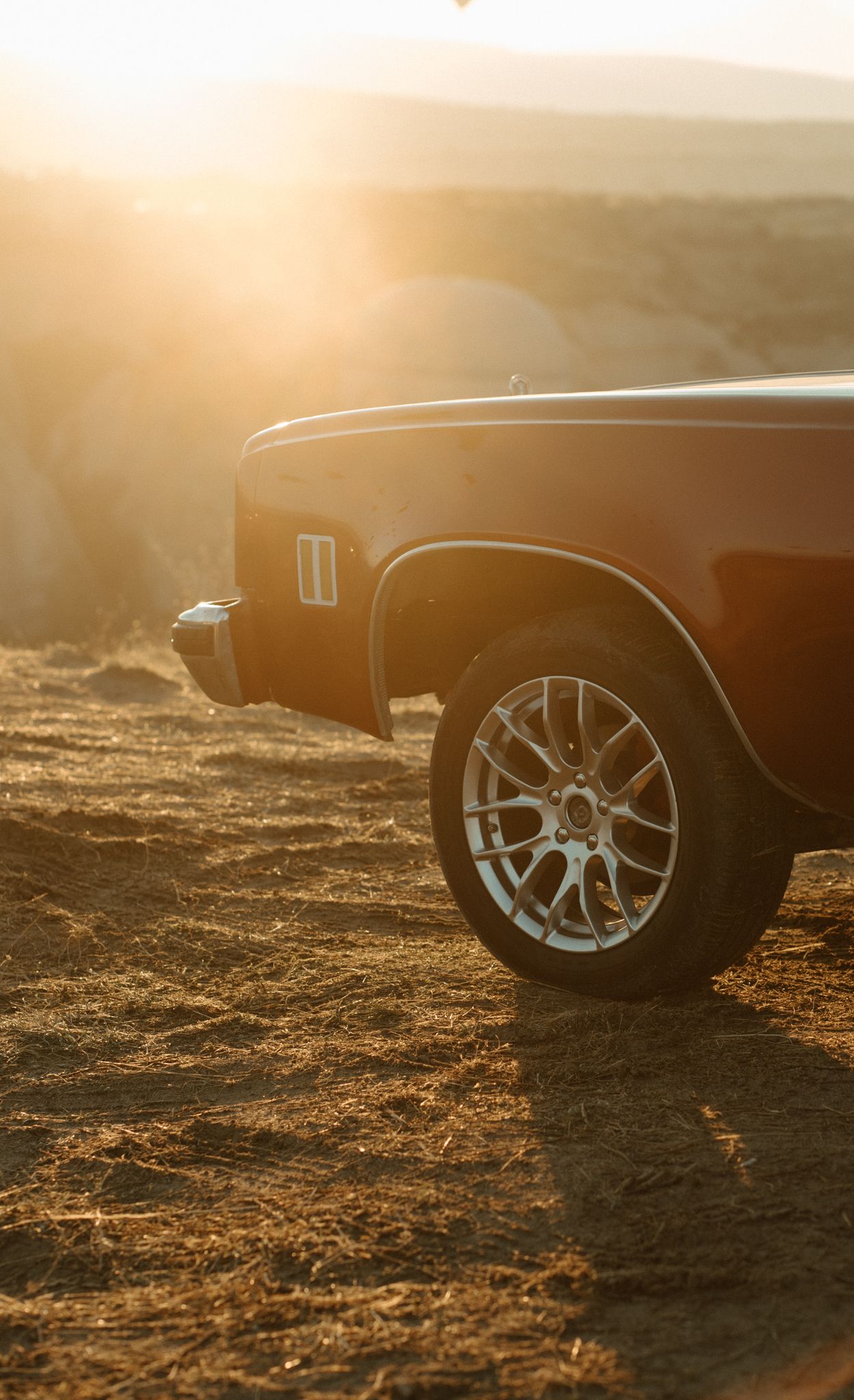 Vintage car wheel and side panel illuminated by golden sunset light on dusty ground.