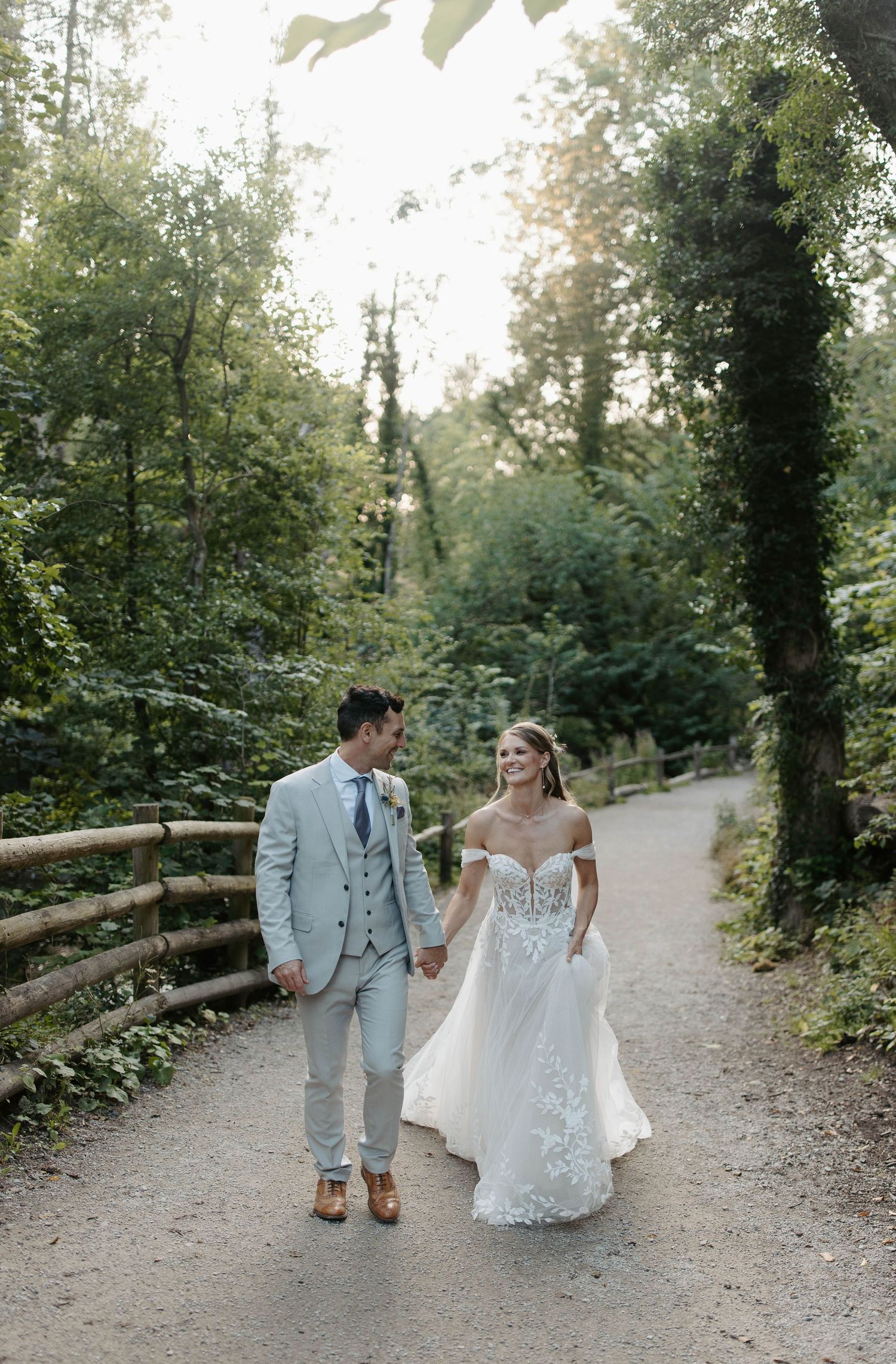Newlyweds walking hand in hand down a tree-lined path in romantic wedding attire.