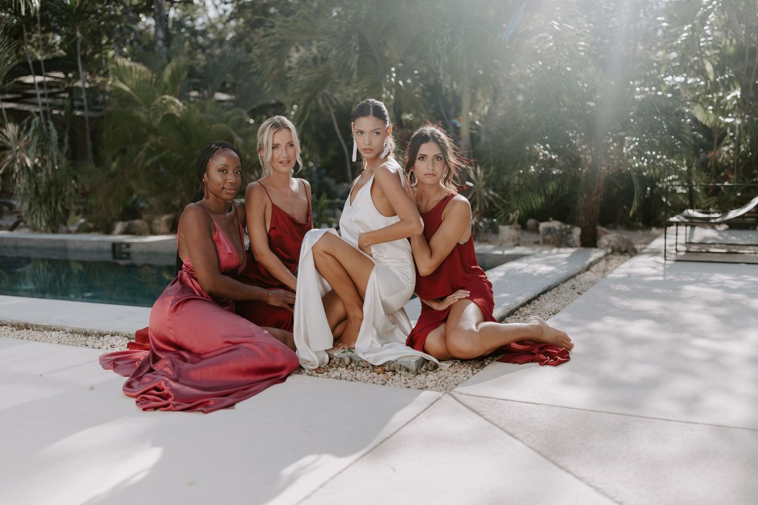 Group of women in flowing red dresses sitting poolside in tropical setting.