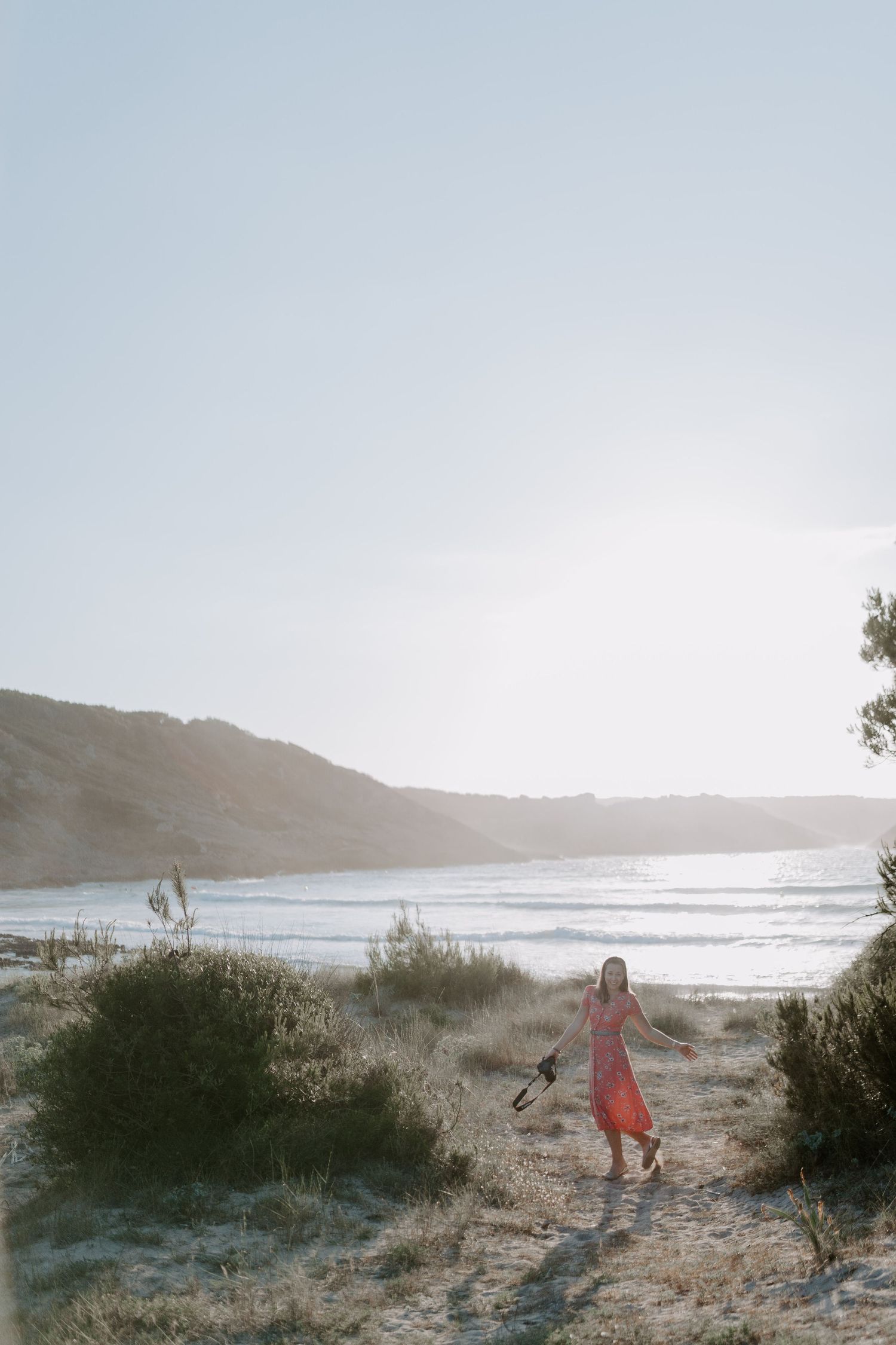 Person in red dress walking through coastal landscape at sunset.