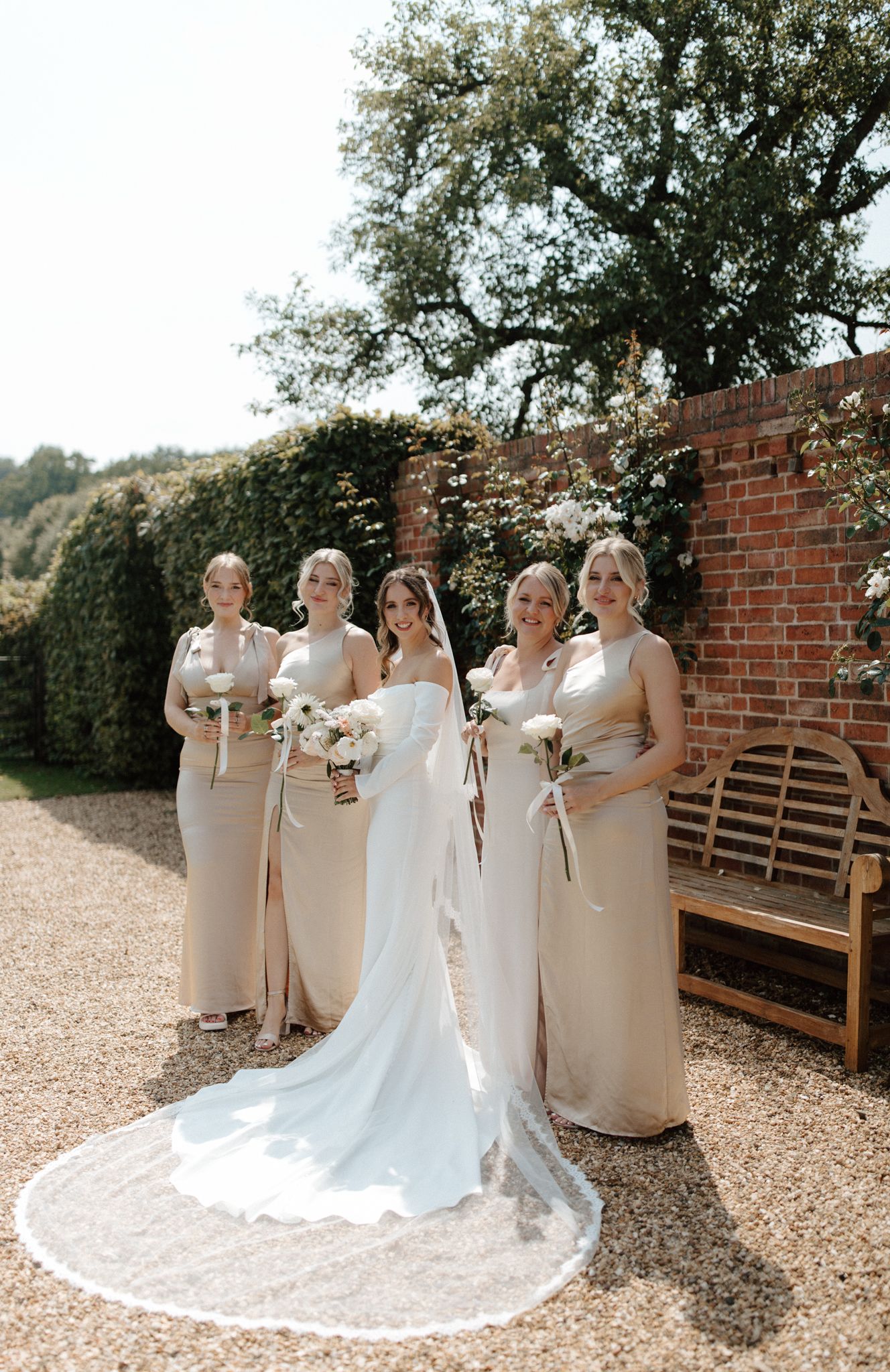 Wedding party in cream-colored dresses pose against a brick wall with a flowing white bridal gown.