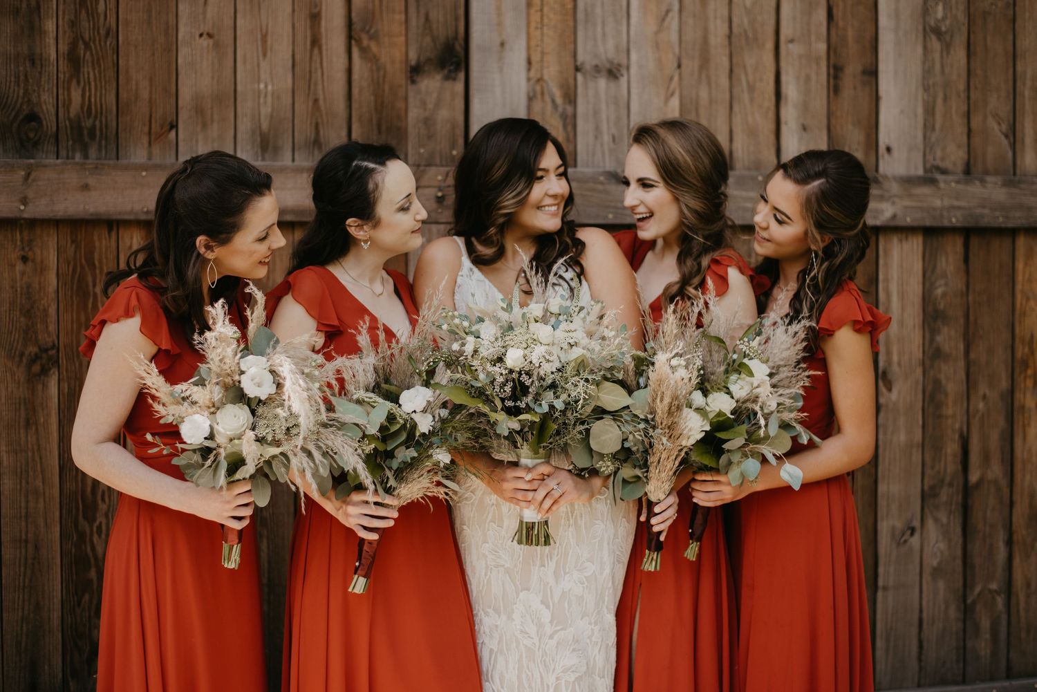 Bride laughing with bridesmaids in burnt orange dresses holding bouquets, photographed by Willow + Vine Photography in a warm, candid m