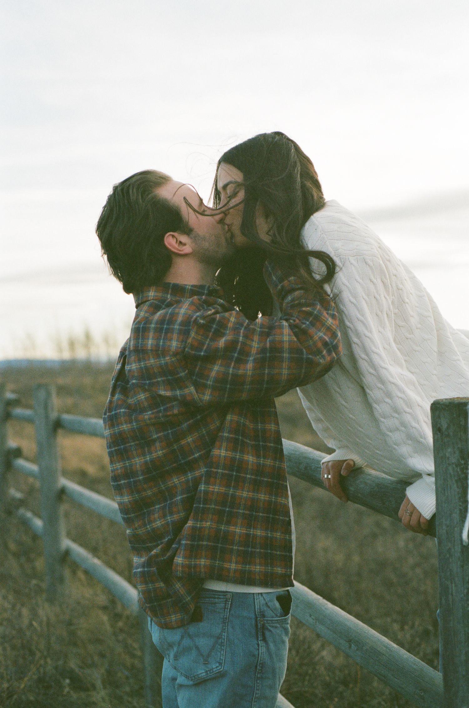 A couple embraces tenderly by a wooden fence post in a rural landscape.