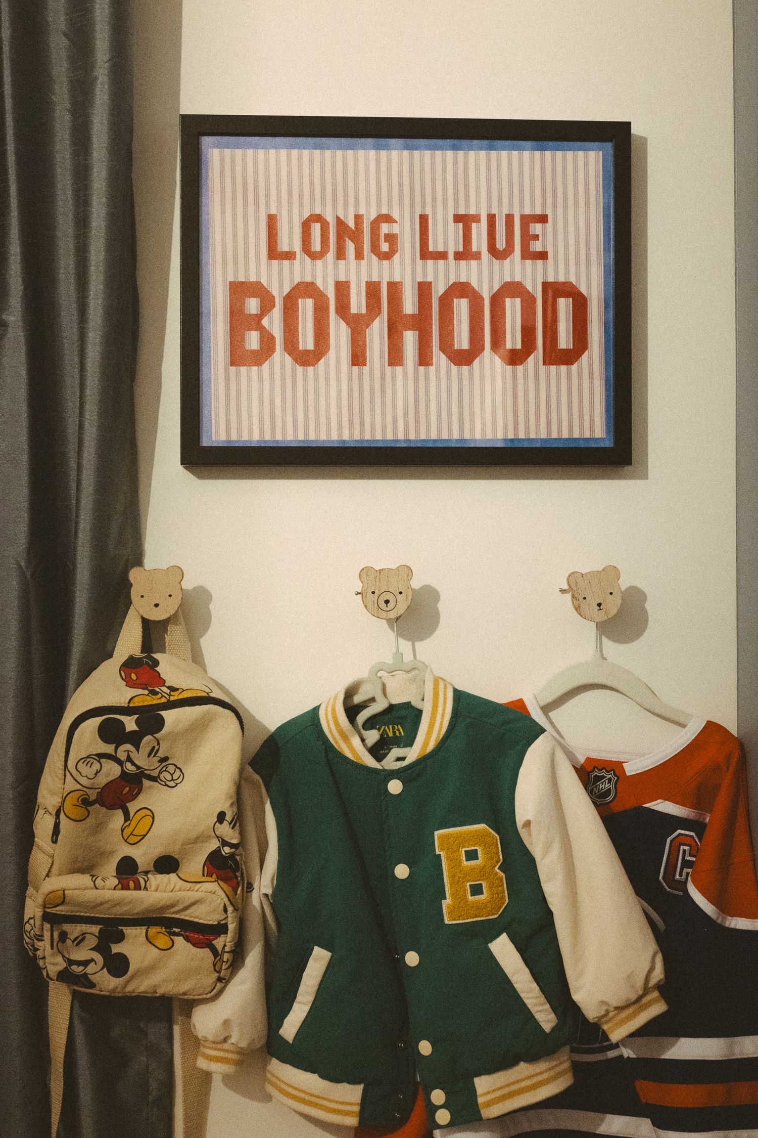 Vintage varsity letterman jackets and sports memorabilia displayed on wall hooks beneath framed text art in a bedroom.