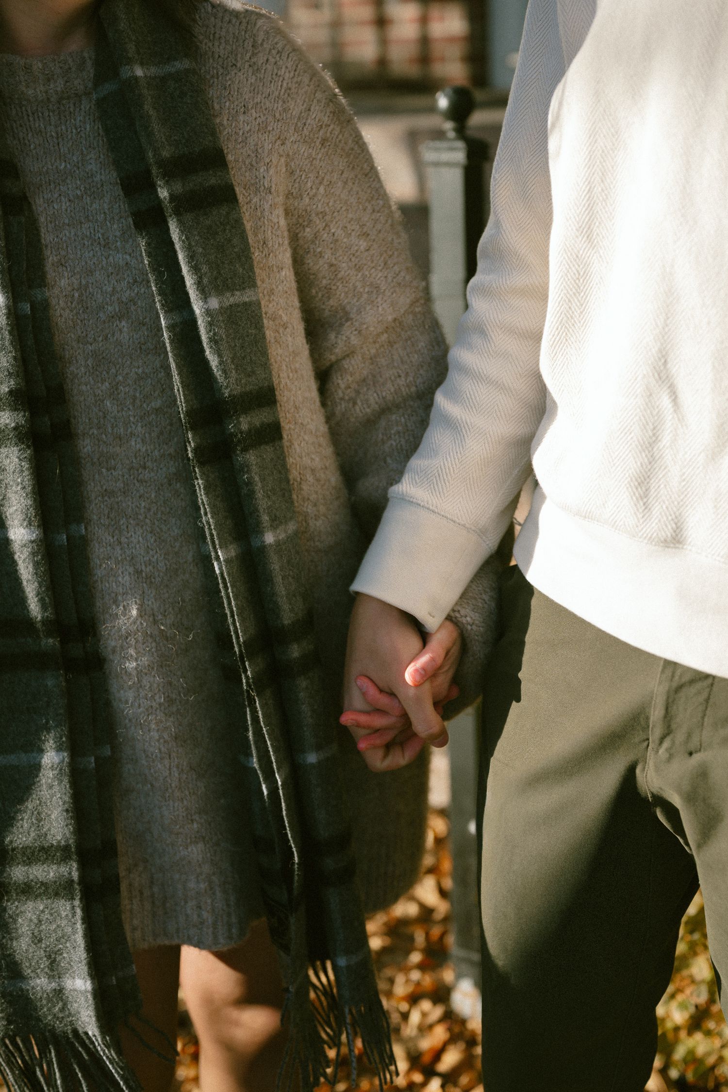 Close-up shot of two people holding hands while wearing contrasting gray and white sweaters with autumn leaves on ground.