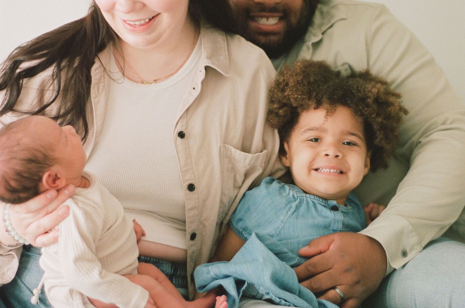 Happy family portrait with parents holding newborn baby and smiling toddler in light, airy studio setting.