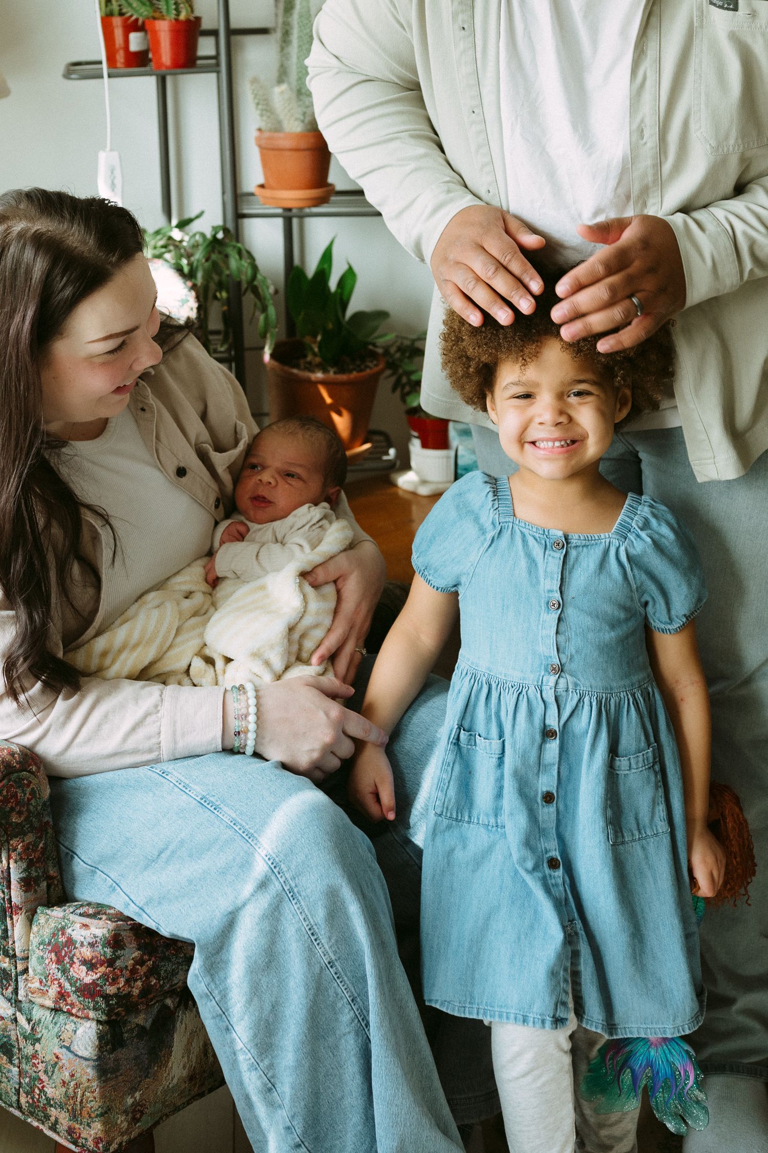 A joyful family poses indoors with a mother holding a newborn, a smiling girl in a blue dress, and plants in the background.