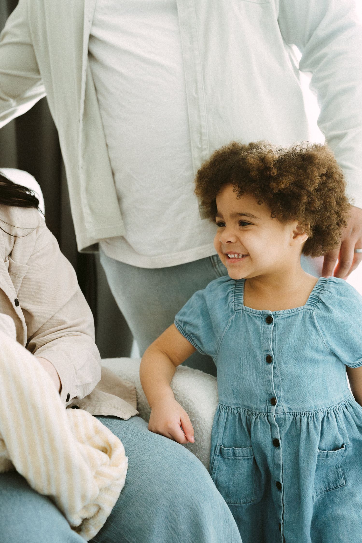 Happy curly-haired toddler girl in denim dress smiling at family member indoors.