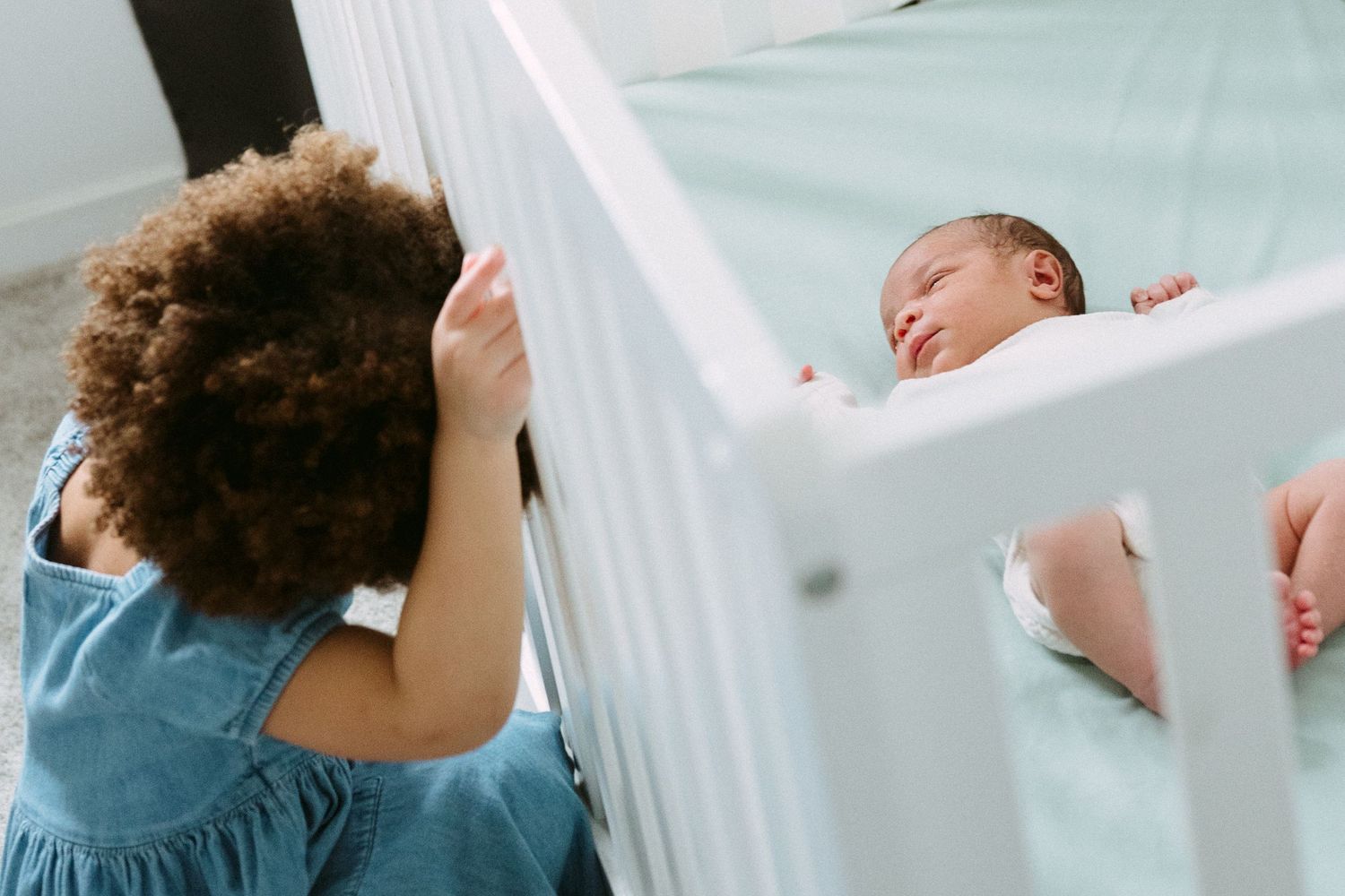 A curly-haired toddler in a blue dress peers through white crib rails at a peacefully sleeping newborn baby.