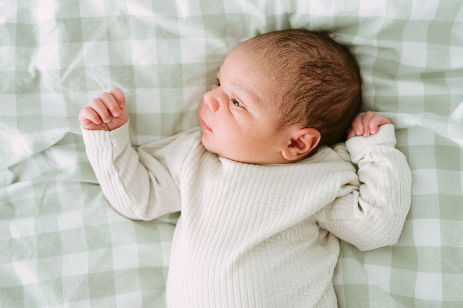 Newborn baby sleeping peacefully on green gingham blanket wearing white ribbed onesie with tiny fist raised.