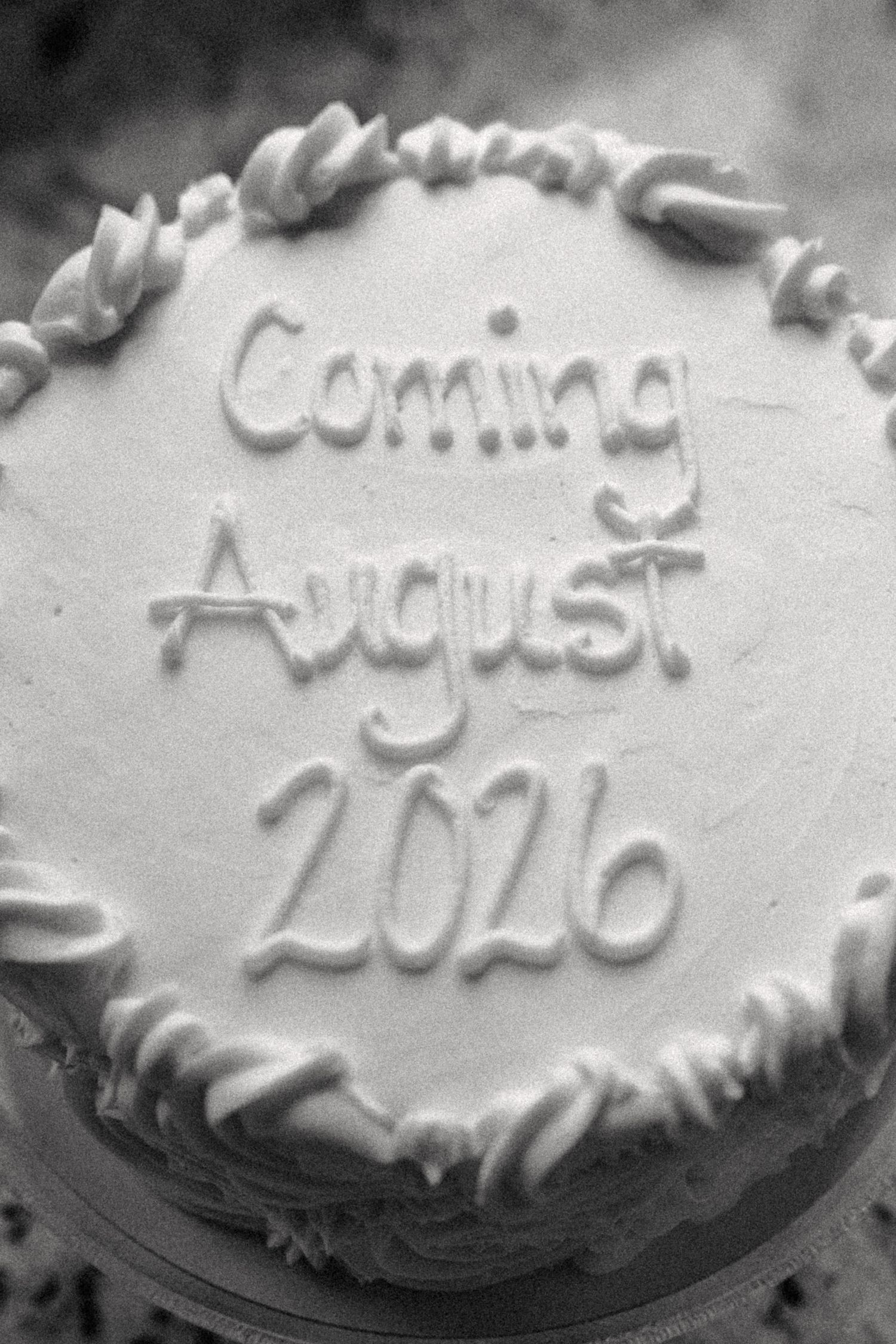 Black and white photo of decorated cake with Coming August 2026 written in frosting for baby announcement.