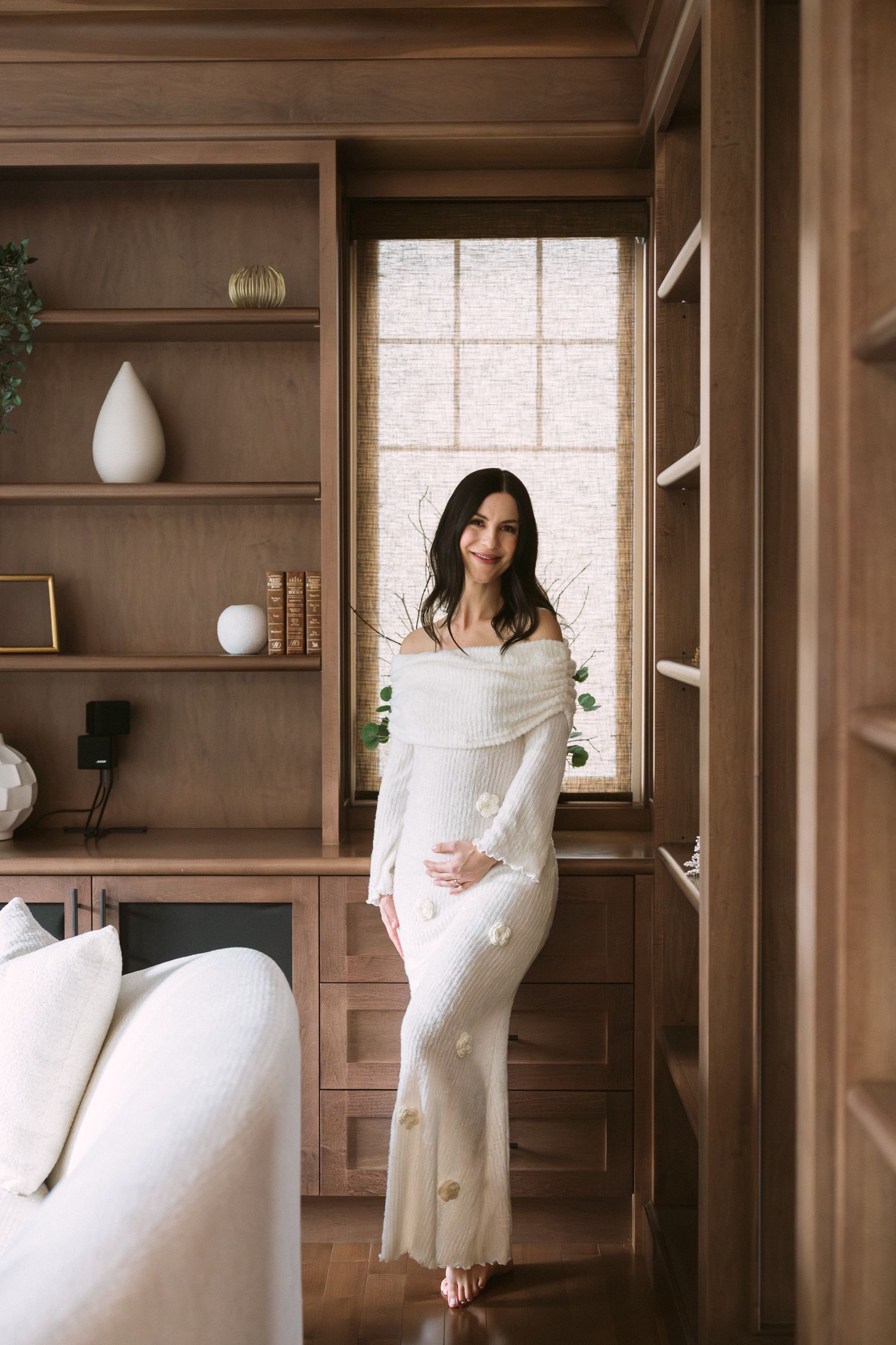 Pregnant woman in elegant white off-shoulder gown standing by window in wood-paneled room.