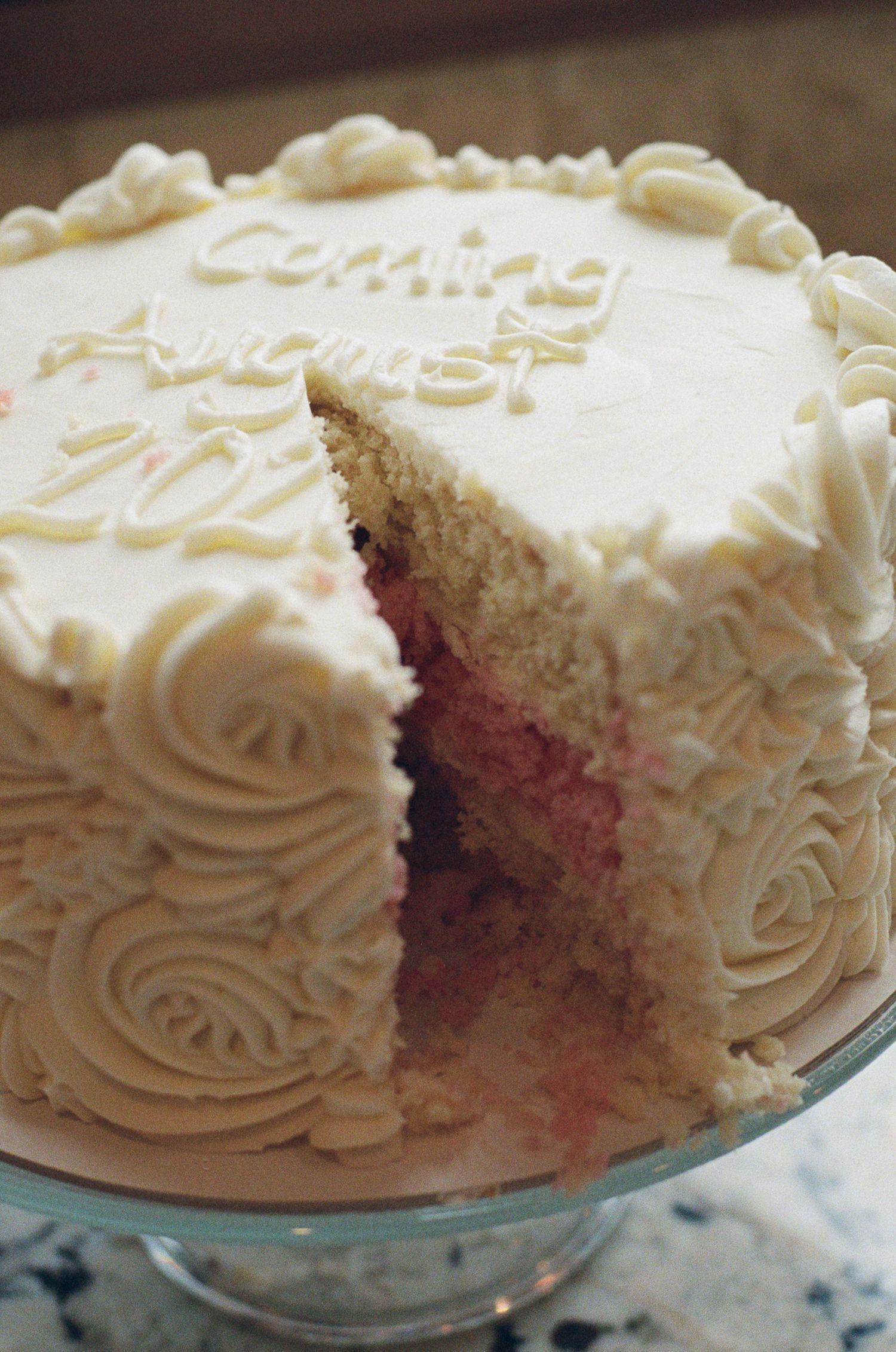 Gender reveal cake with pink interior and white frosting rosettes reading Coming August 2026 on cake stand.