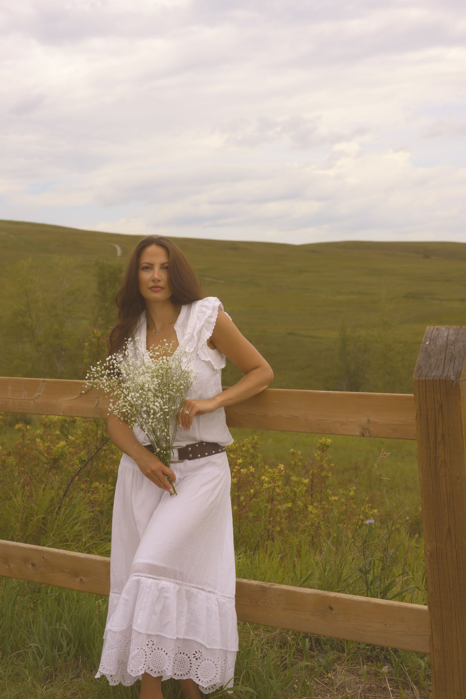 Person in white eyelet dress stands by rustic fence holding fresh wildflowers.