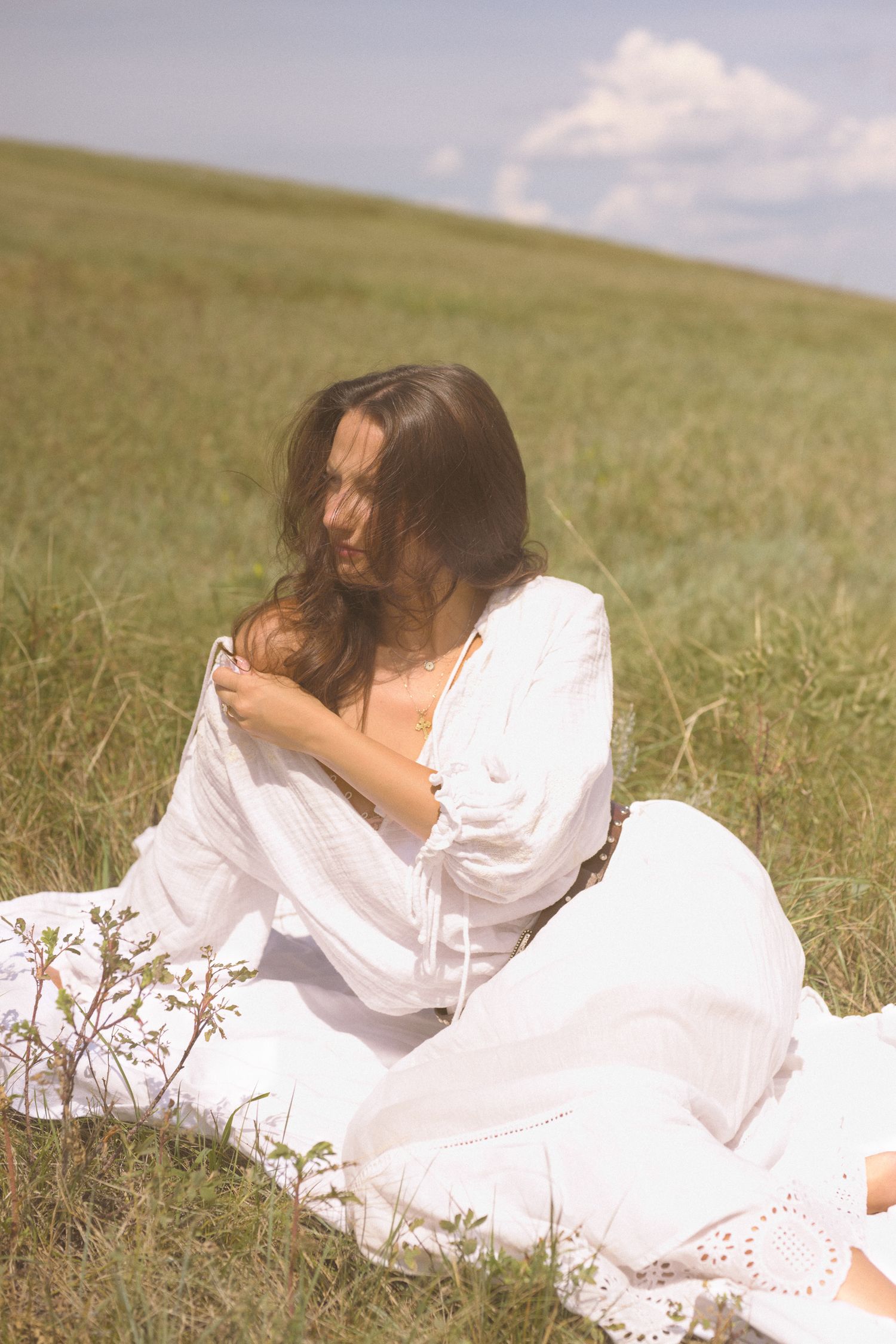 A figure in a flowing white dress sits gracefully in a sunlit meadow on a summer day.