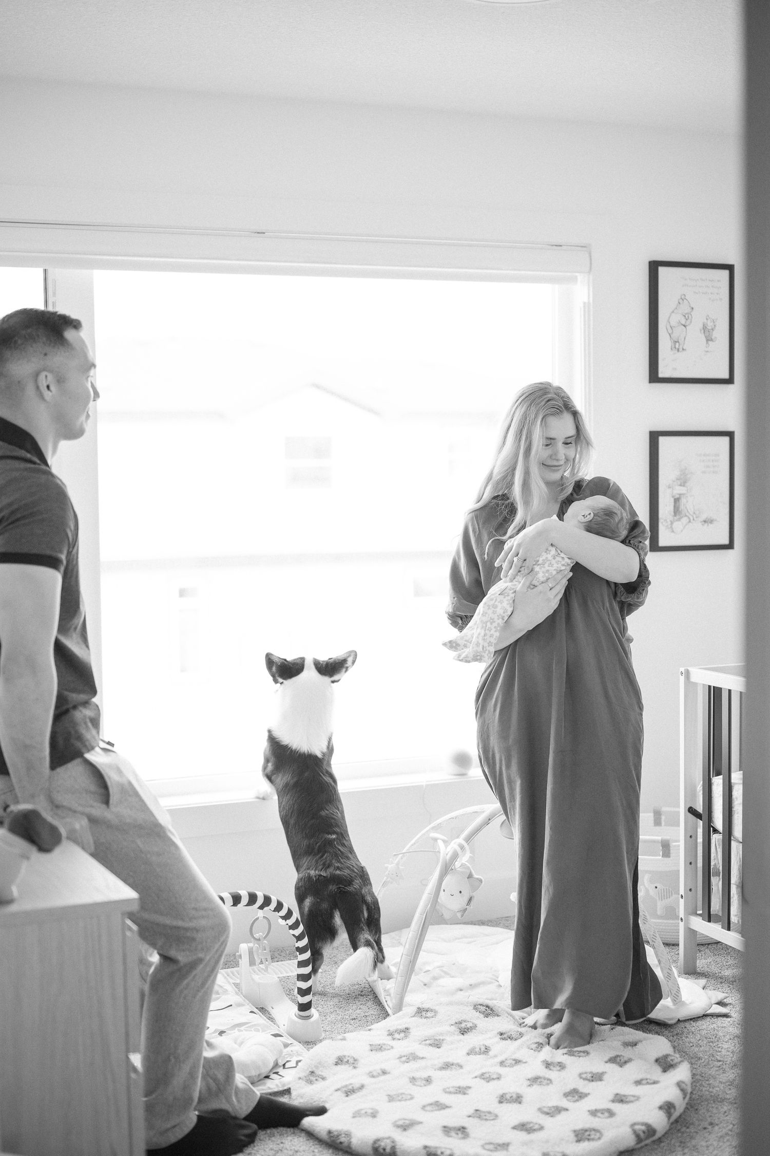 A couple stands in a bright nursery with their baby while their cat playfully jumps in the foreground.