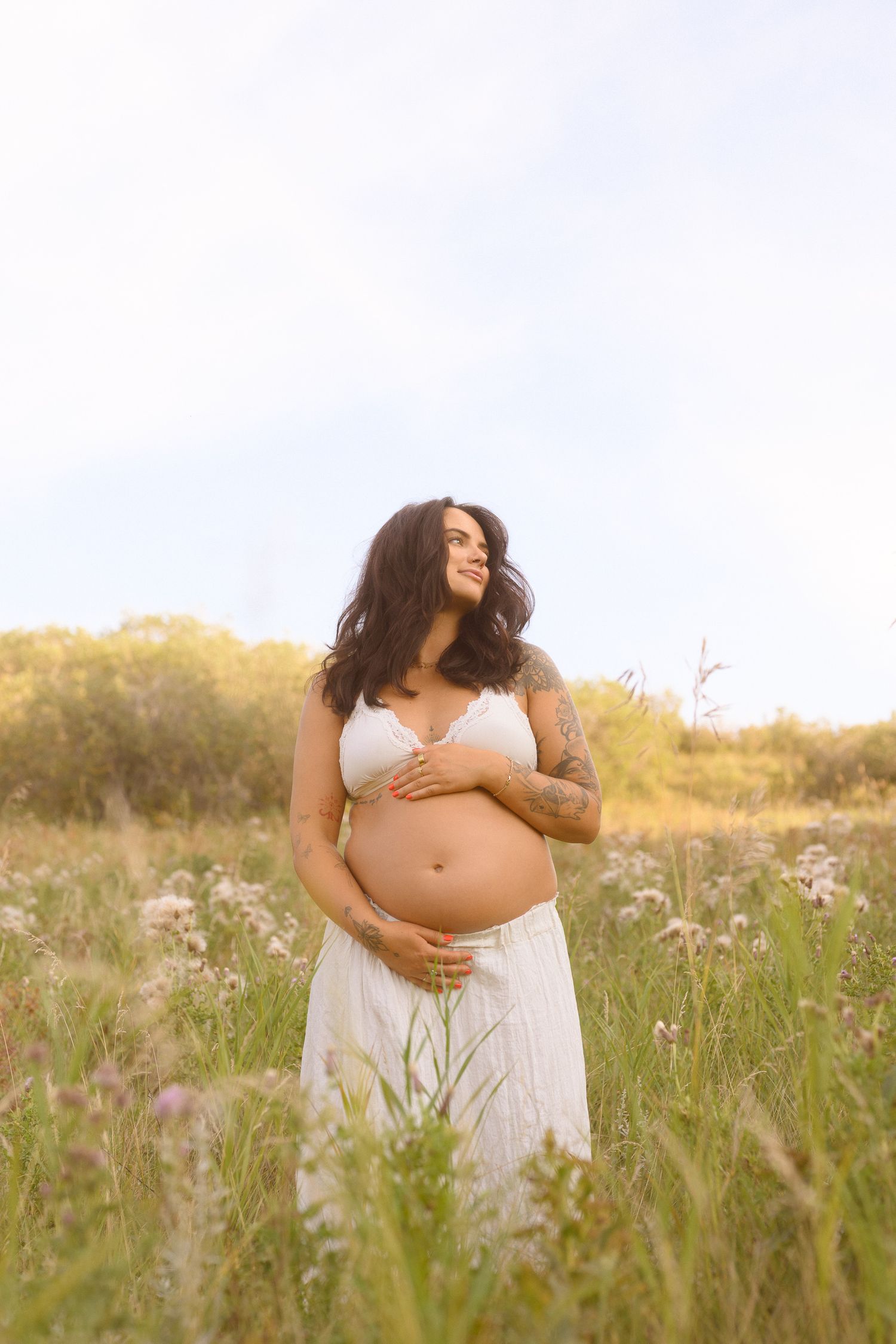 A maternity photoshoot in a sunlit meadow featuring a flowing white dress and gentle embraces.