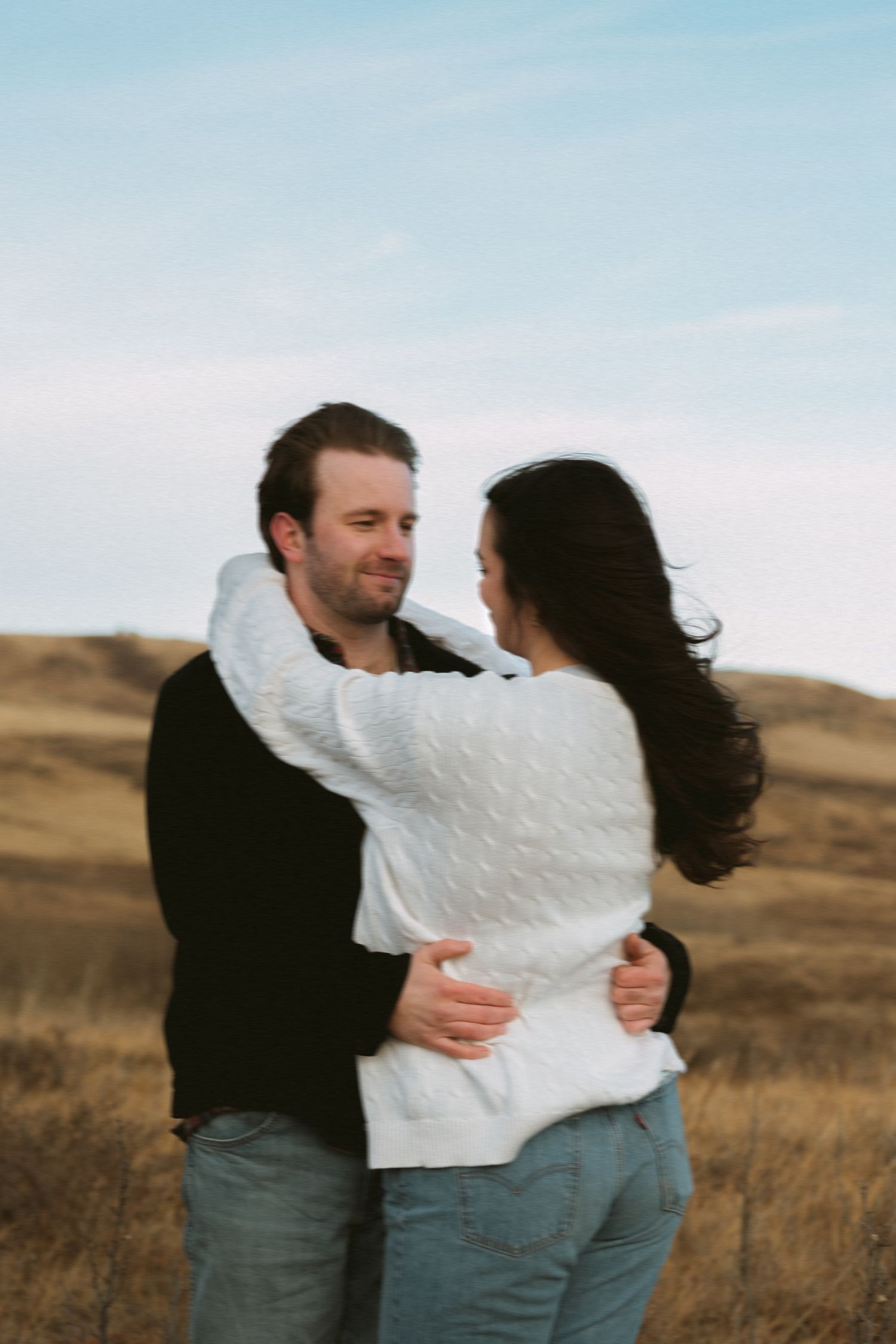 Couple embracing in an open field against a blue sky backdrop.